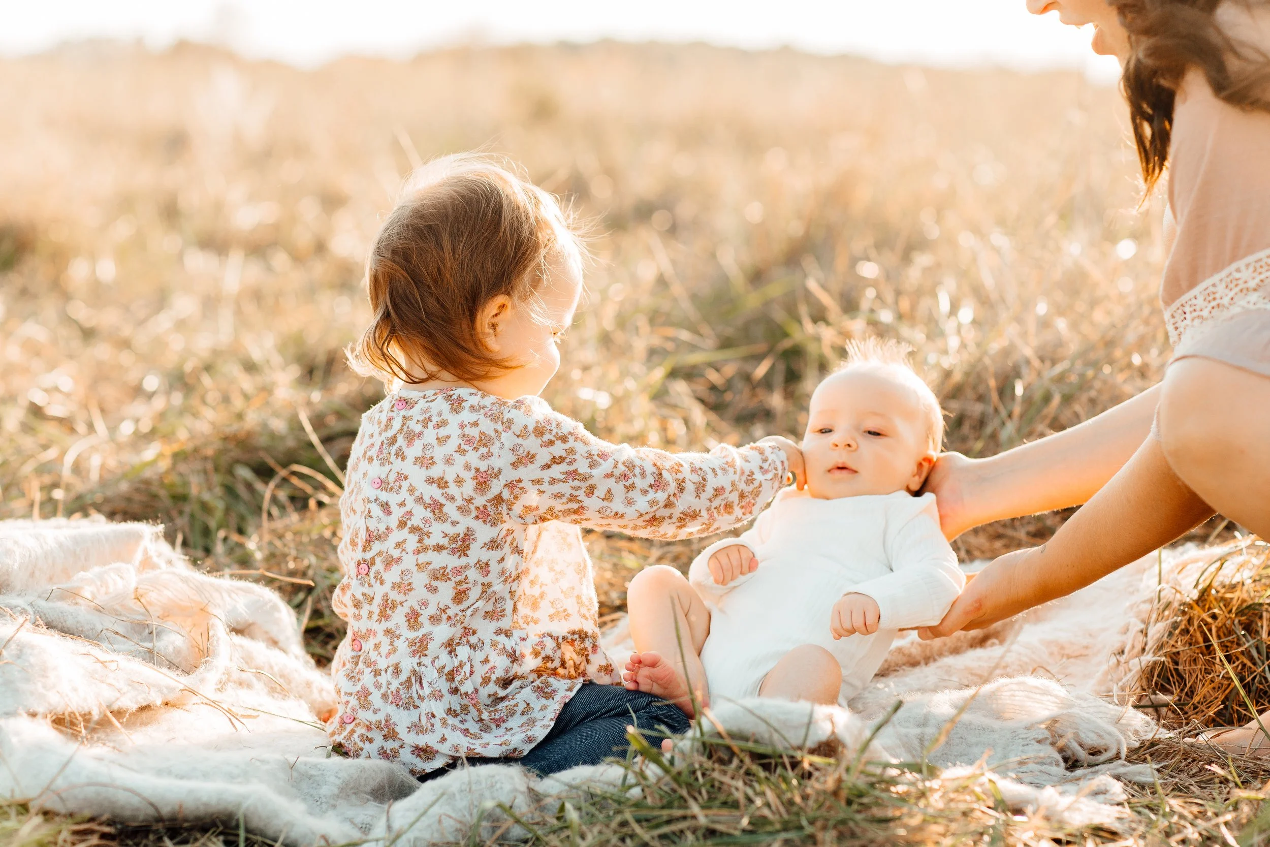 A young girl and her mother playing with a baby on a blanket in a field during golden hour.