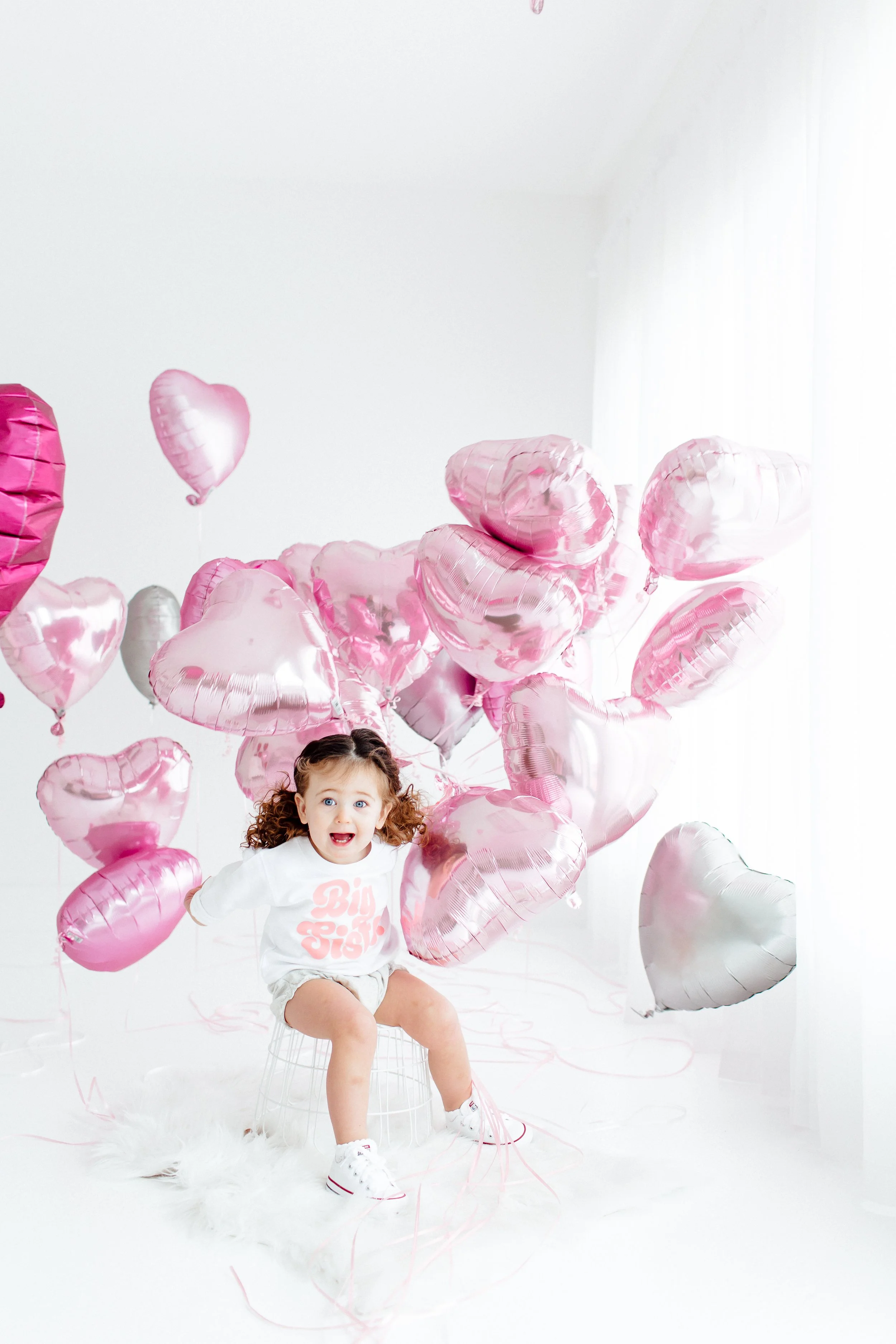 A young girl with curly hair sitting on a stool surrounded by pink and silver heart-shaped balloons in a white room.
