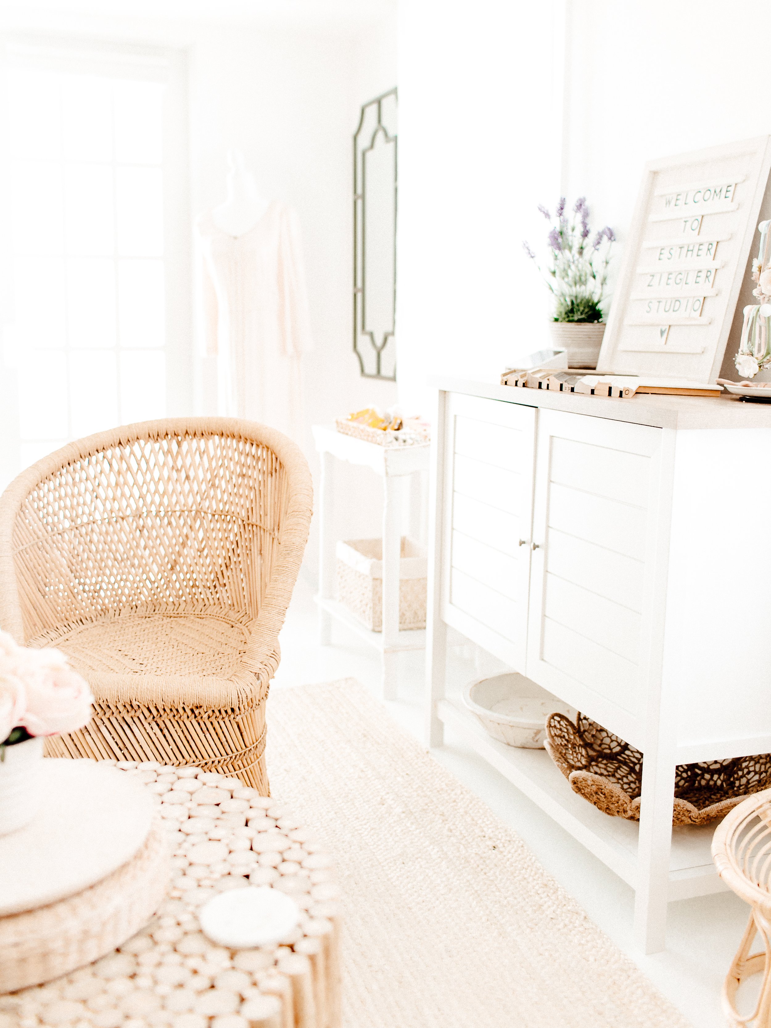 A bright, airy room with neutral decor, featuring a wicker chair, a white cabinet with baskets, a sign that says "Welcome to Esther Ziegler Studio," and a plant with purple flowers.