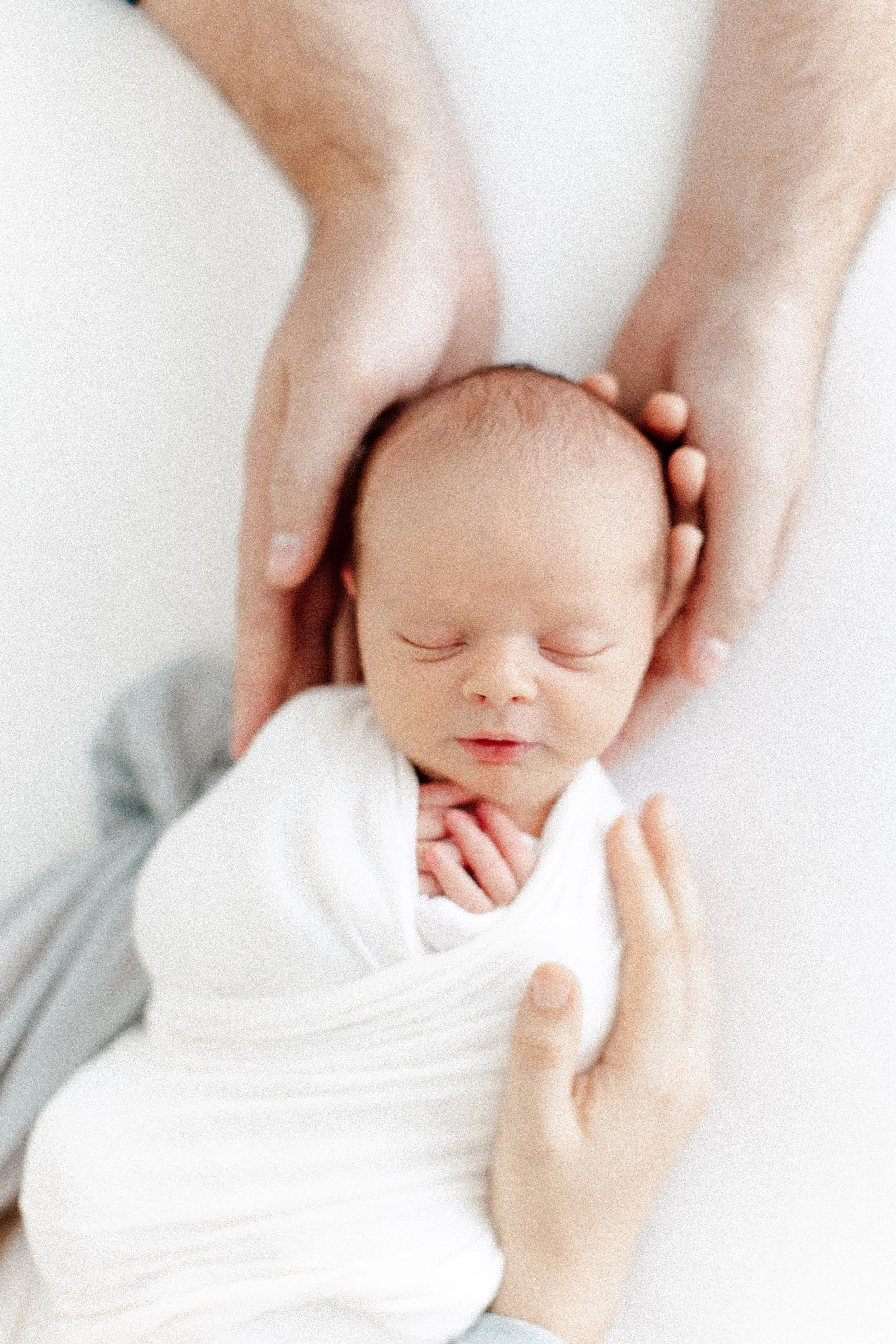 Close-up of a newborn baby wrapped in a white blanket, sleeping peacefully with two hands gently cradling its head and body.