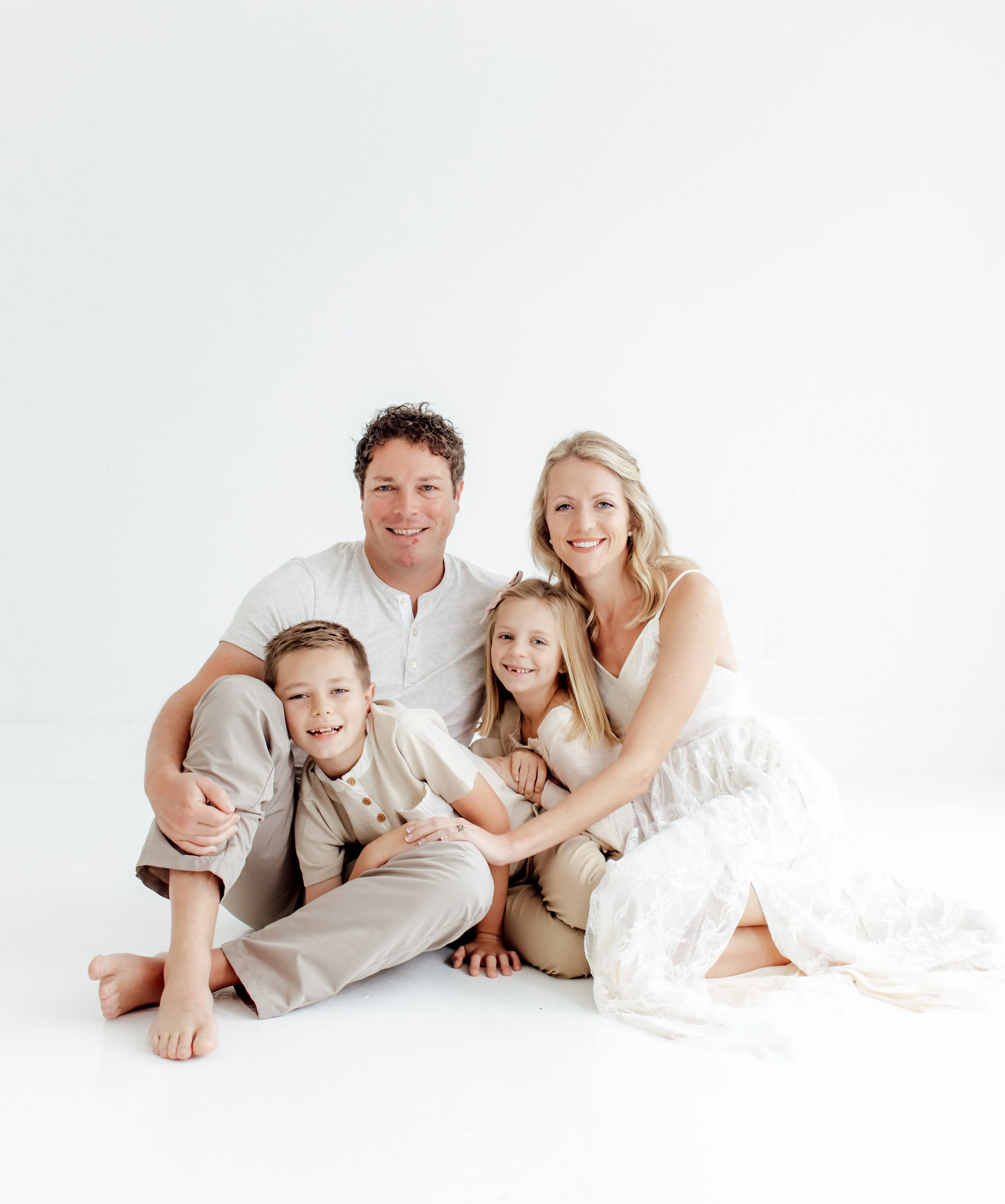 A happy family of four sitting on a white background, smiling at the camera. The family includes a mother, father, son, and daughter, all dressed in light-colored clothing.