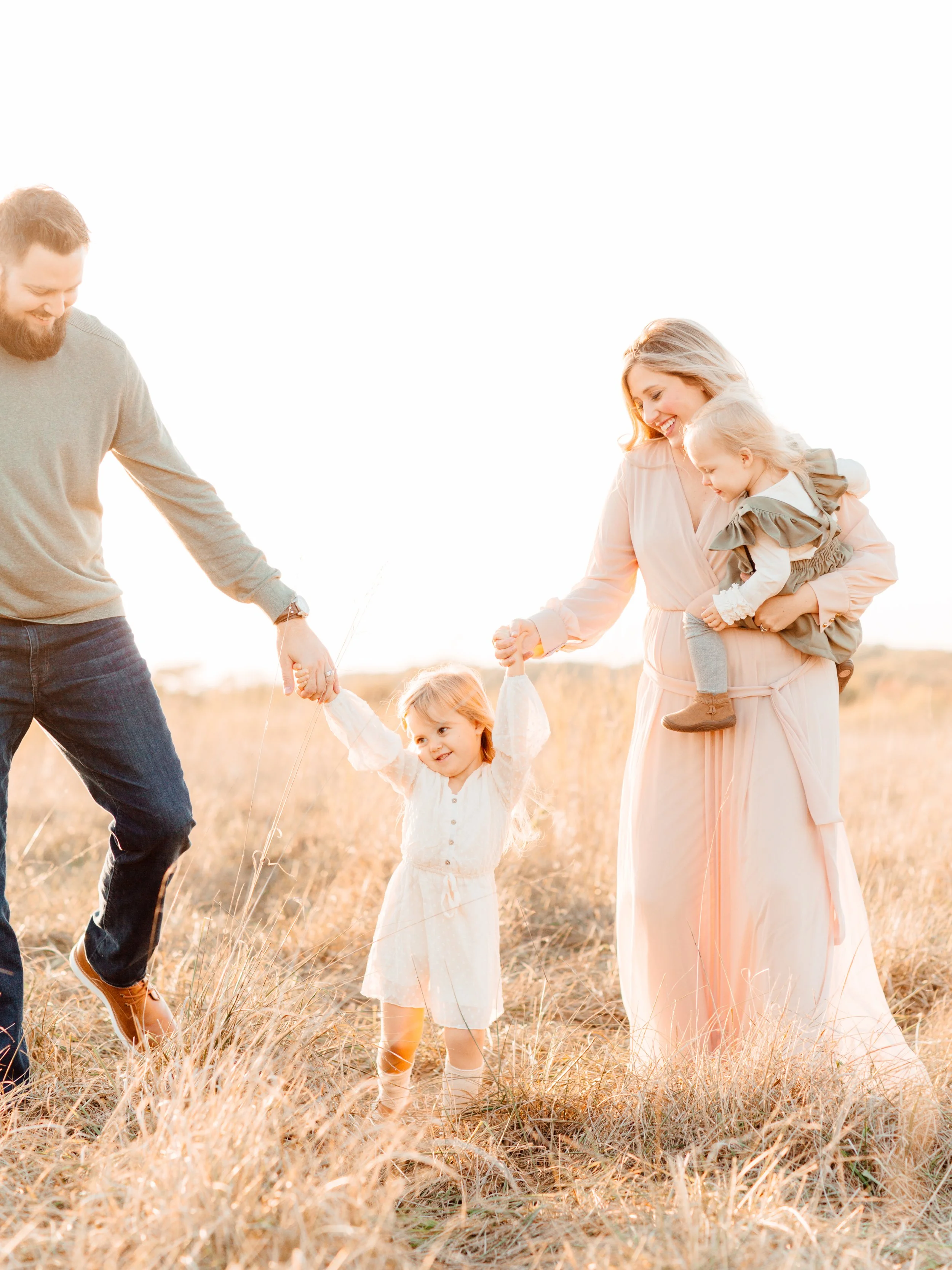A family of four in a grassy field during sunset, holding hands and playing. The father wears a gray sweater and jeans, the mother in a light pink dress holding a young girl in gray-purple ruffled dress, the other girl in a white dress with a bow, sm