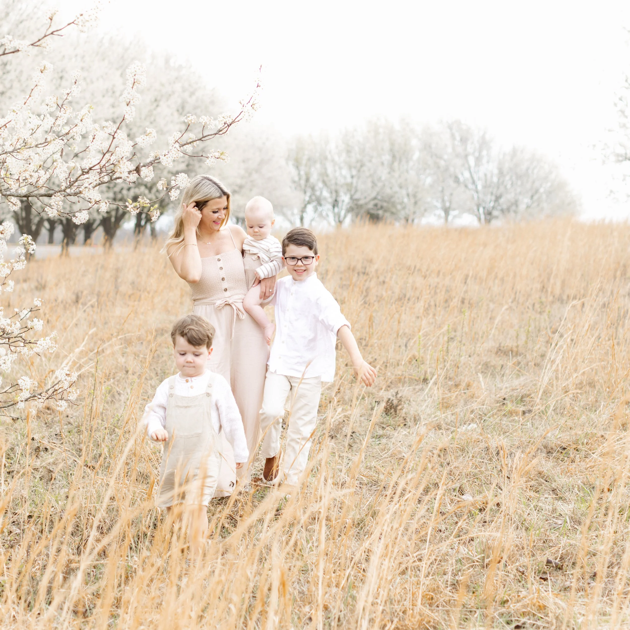 A woman with three young children walking through a golden field with blossoming trees in the background.