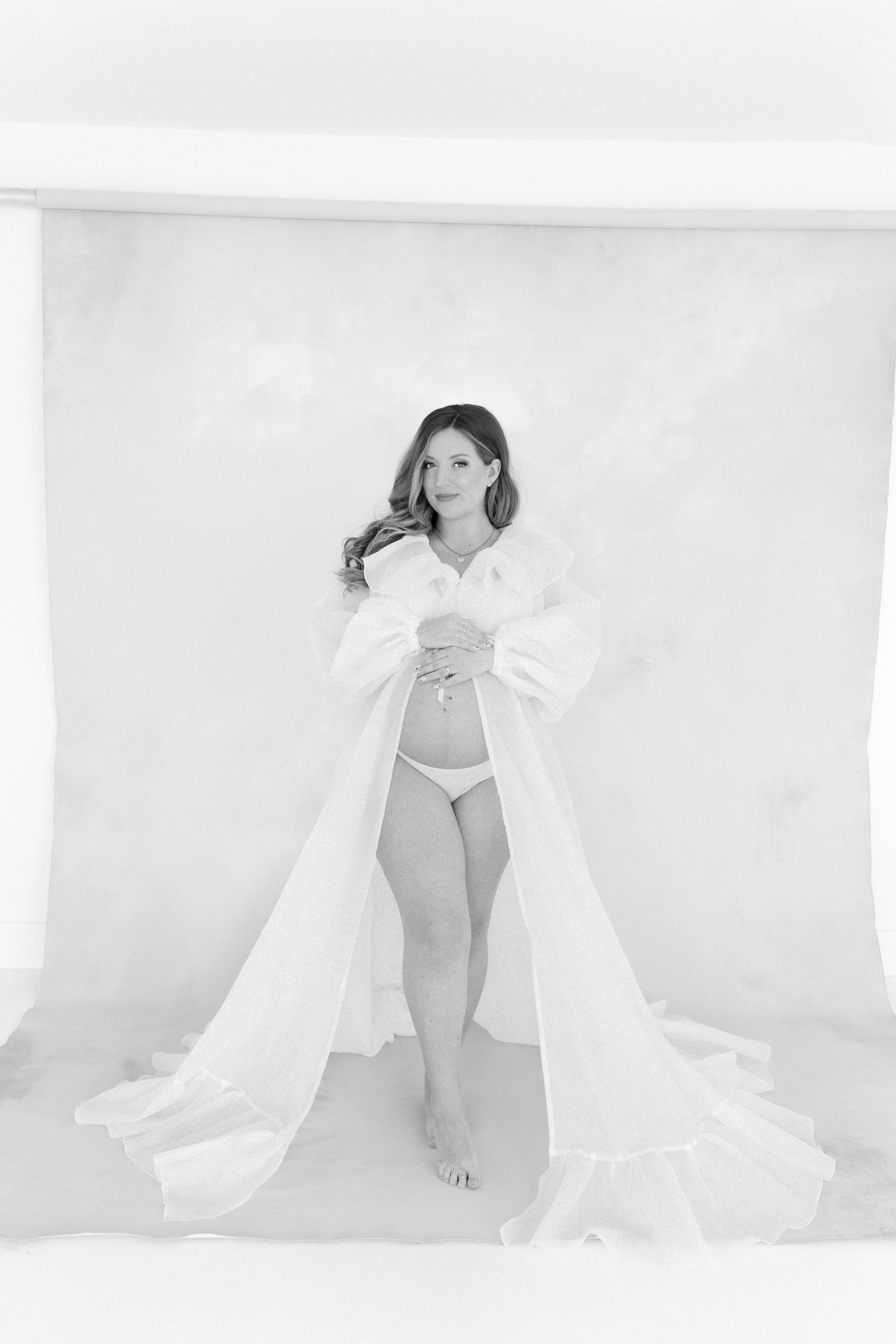 A woman with long wavy hair standing barefoot in front of a plain backdrop, wearing a long, flowing white robe and white underwear, looking at the camera with a slight smile.