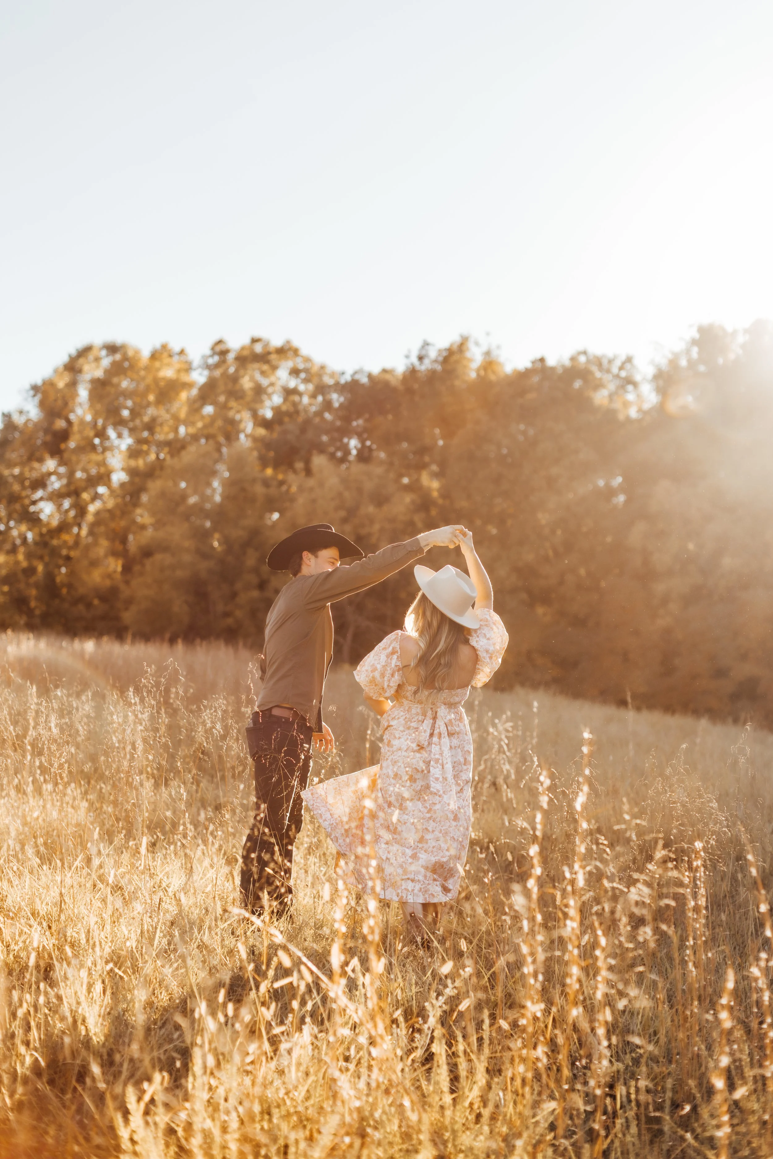 A couple dancing in a field during sunset, with the man wearing a cowboy hat and the woman wearing a large white hat and a floral dress.
