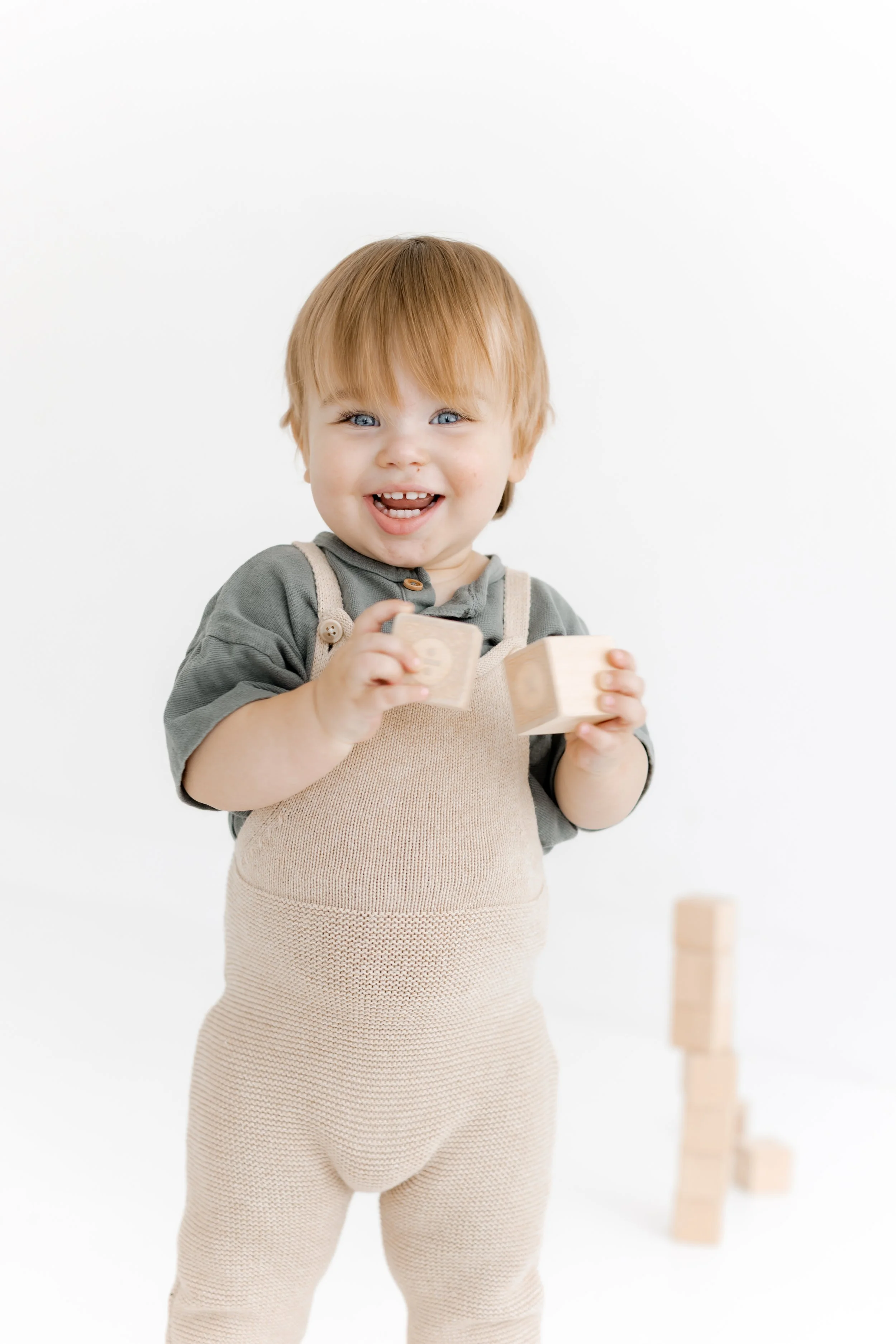 A young boy with red hair and blue eyes smiling and holding wooden blocks, standing in front of a white background with a small stack of blocks.