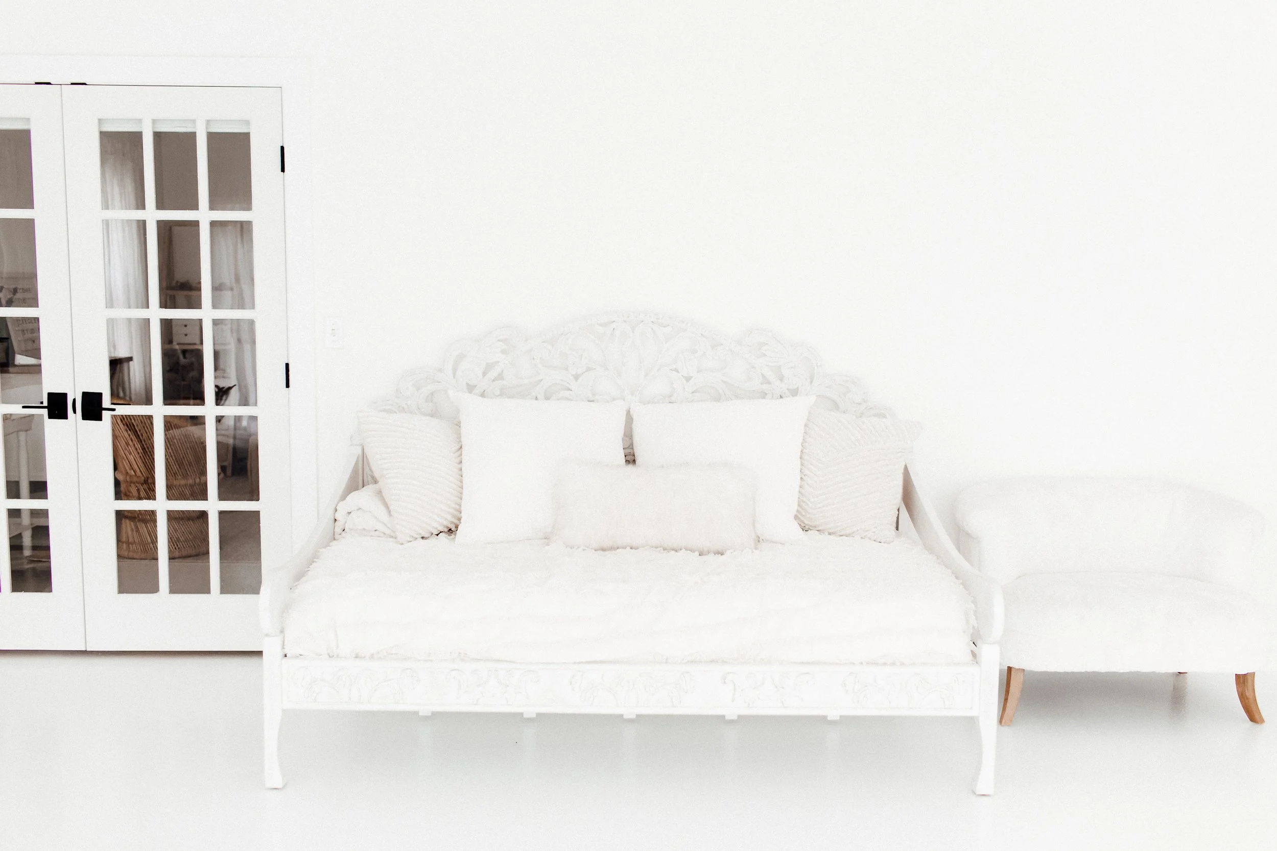 A white vintage style sofa with decorative pillows, next to a white armchair, in a bright room with white walls and a glass-paneled door.