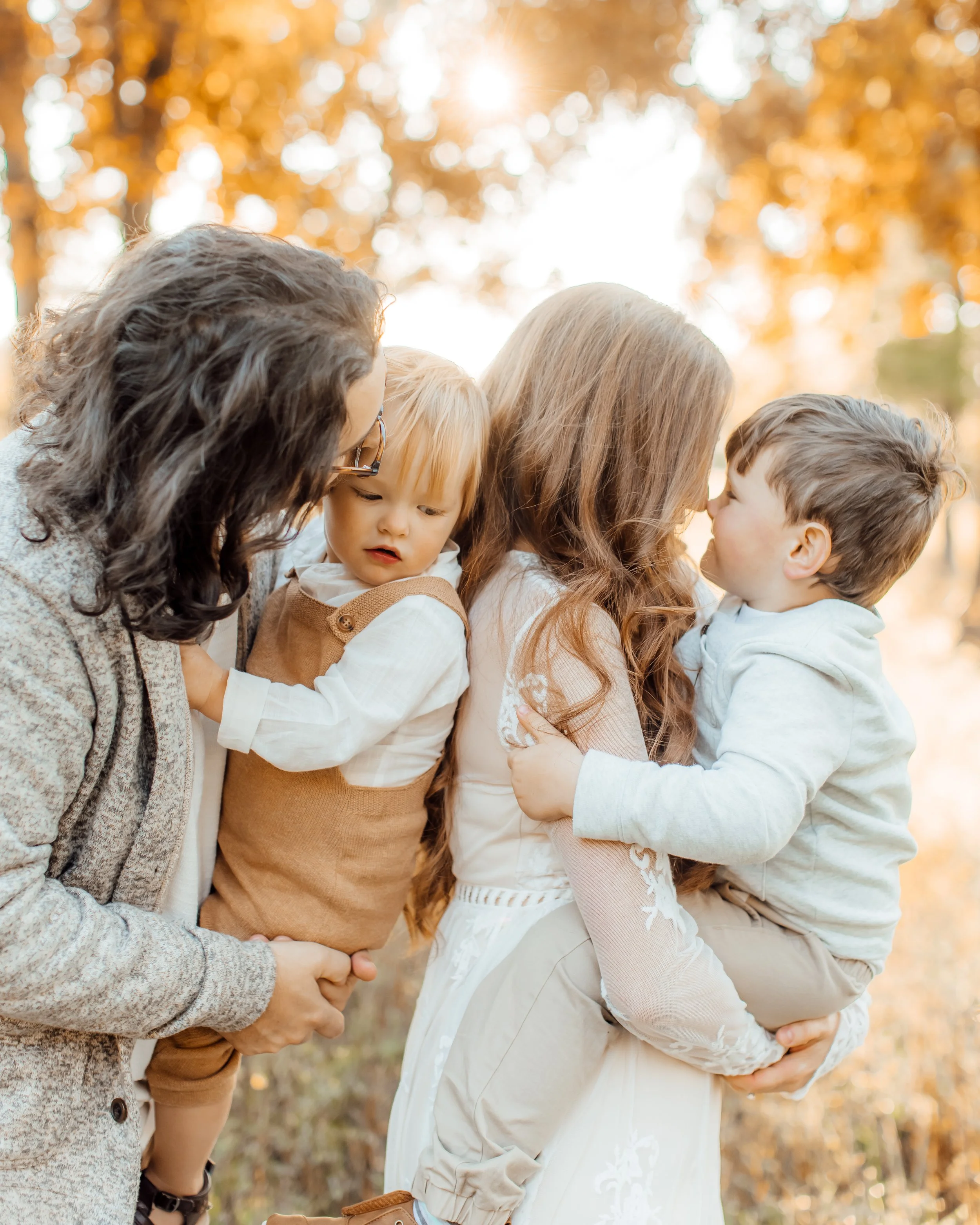 Four individuals, two women and two children, sharing a tender moment outdoors during autumn with warm light and fall foliage in the background.