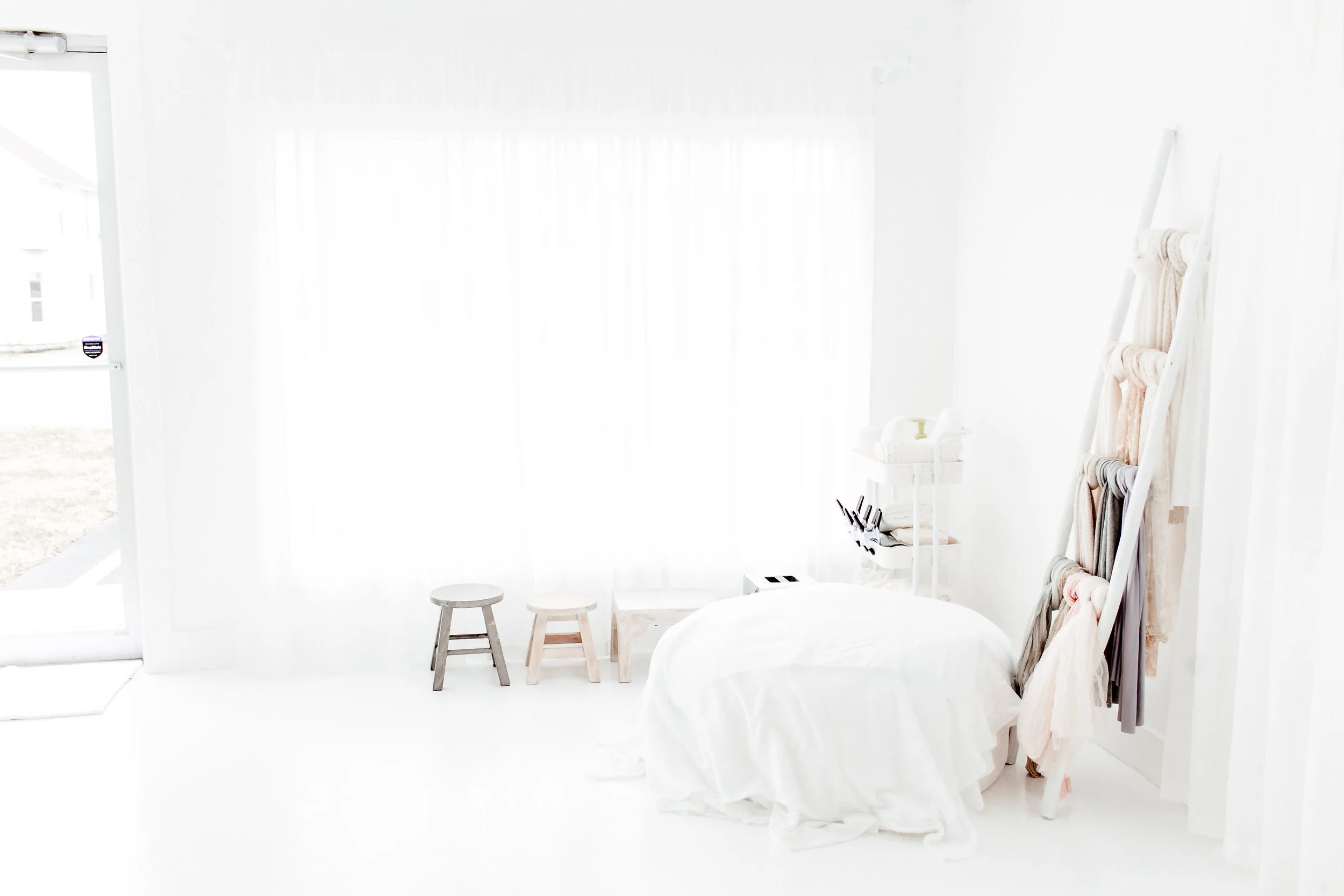 Minimalist white bedroom with bed covered in white linens, wooden stools, a small white shelving unit with towels, a ladder with scarves, and a large window with sheer curtains.
