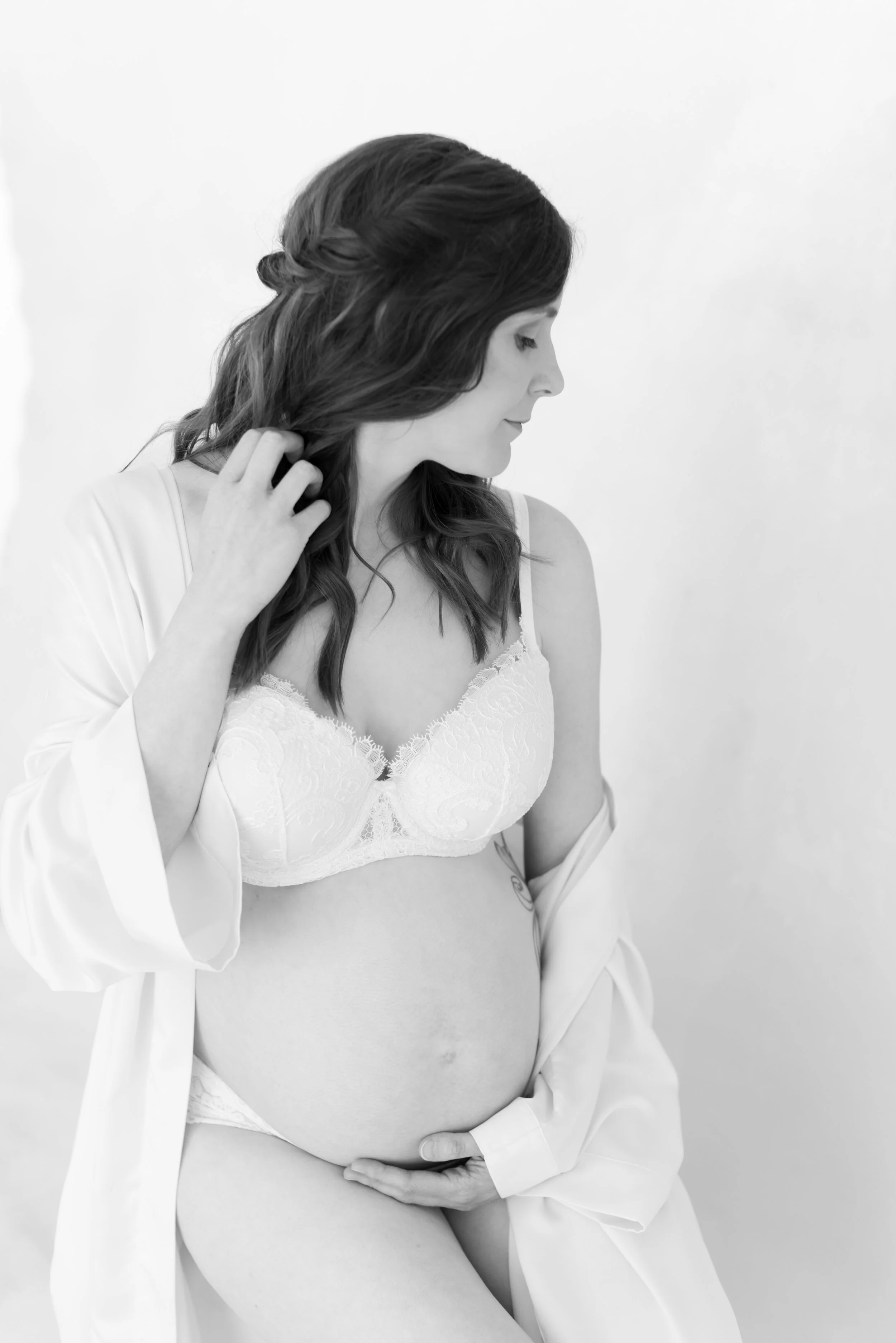 Black and white photo of a pregnant woman in lingerie, gently touching her belly and looking down.