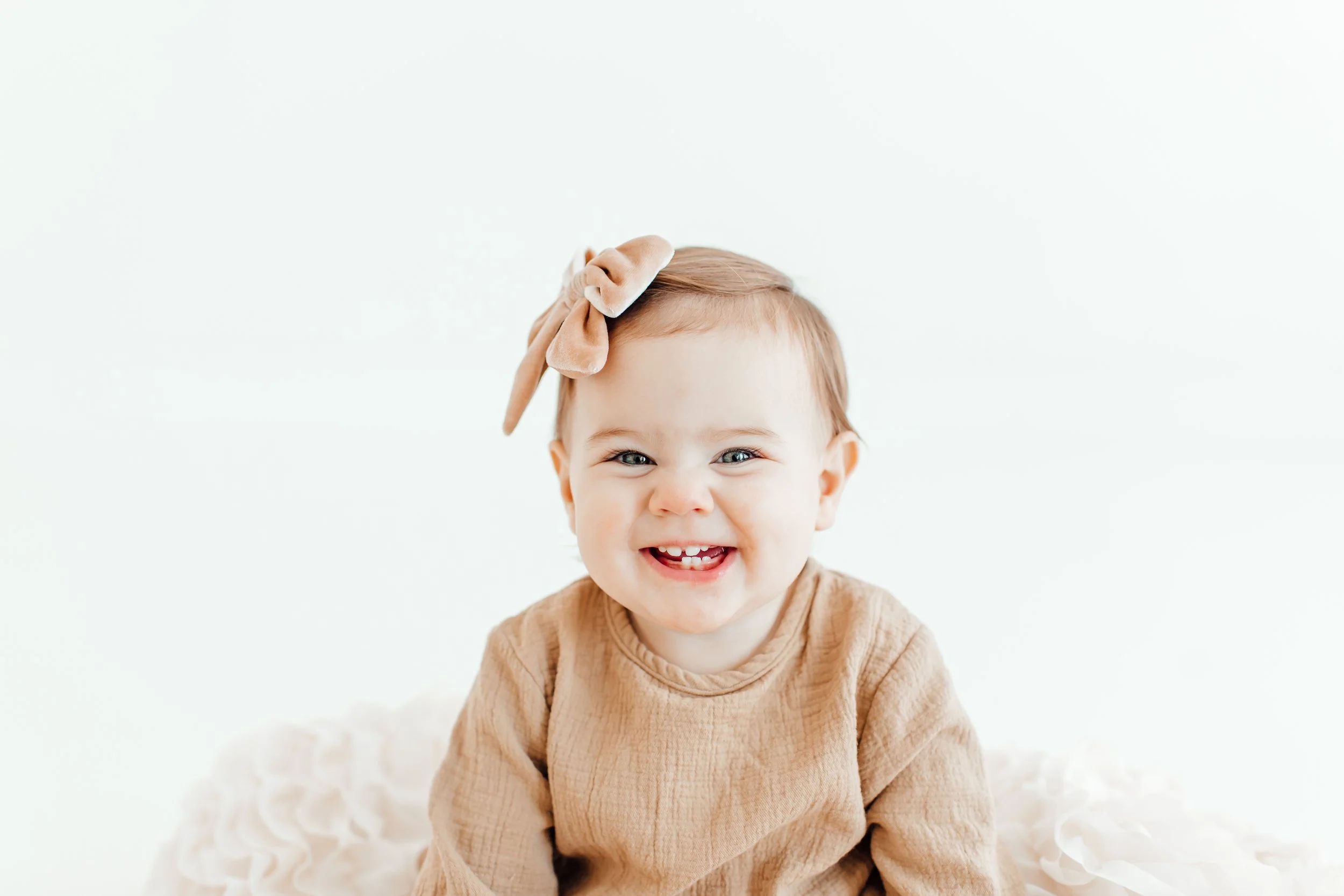 Smiling baby girl with a beige bow on her head, wearing a beige long-sleeved top, sitting on a soft white surface against a white background.