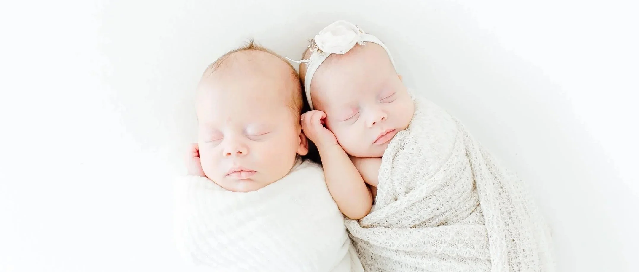 Two sleeping newborn babies, one wrapped in a white blanket and the other in a lace blanket, with one wearing a headband with a flower, laying close together on a white background.