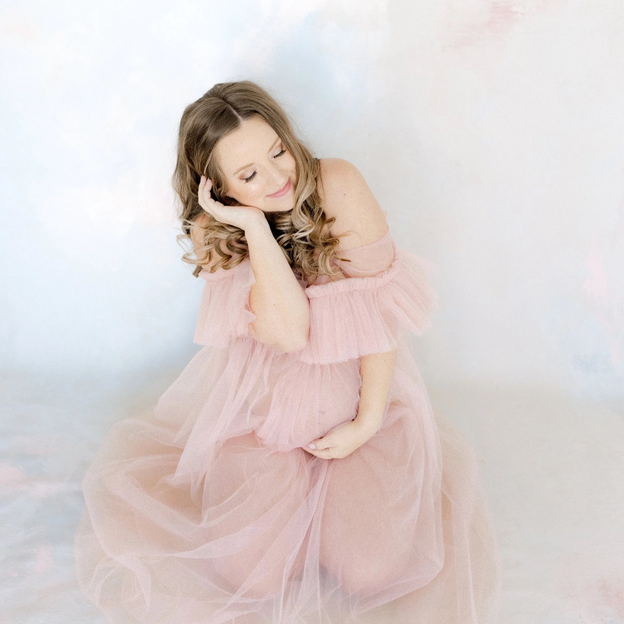 A woman in a pink dress sitting on the floor, with her eyes closed and a gentle smile, touching her head with one hand and resting the other hand on her knee.