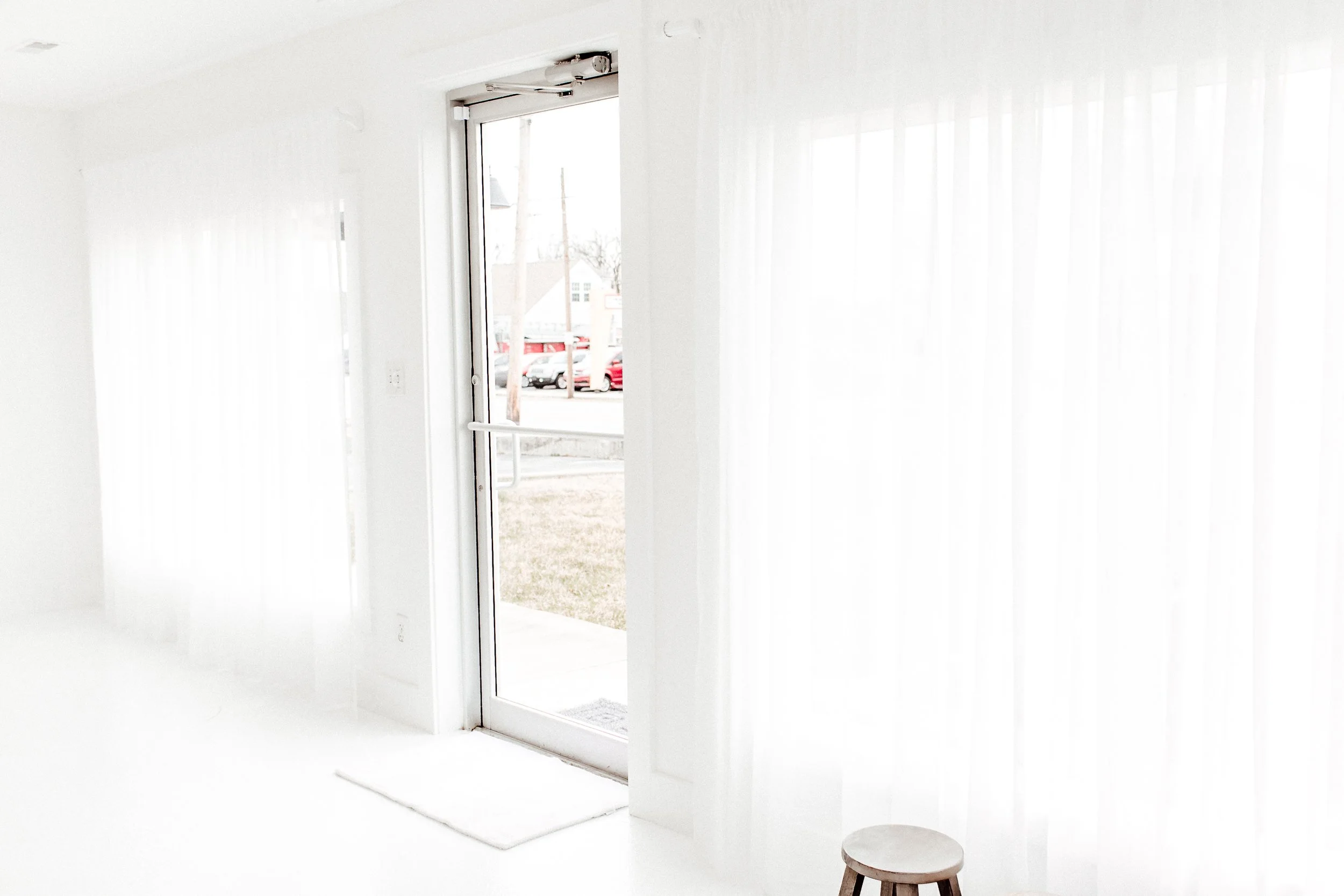 Bright white room with sheer curtains, glass door, small round stool, and view of parked cars outside.