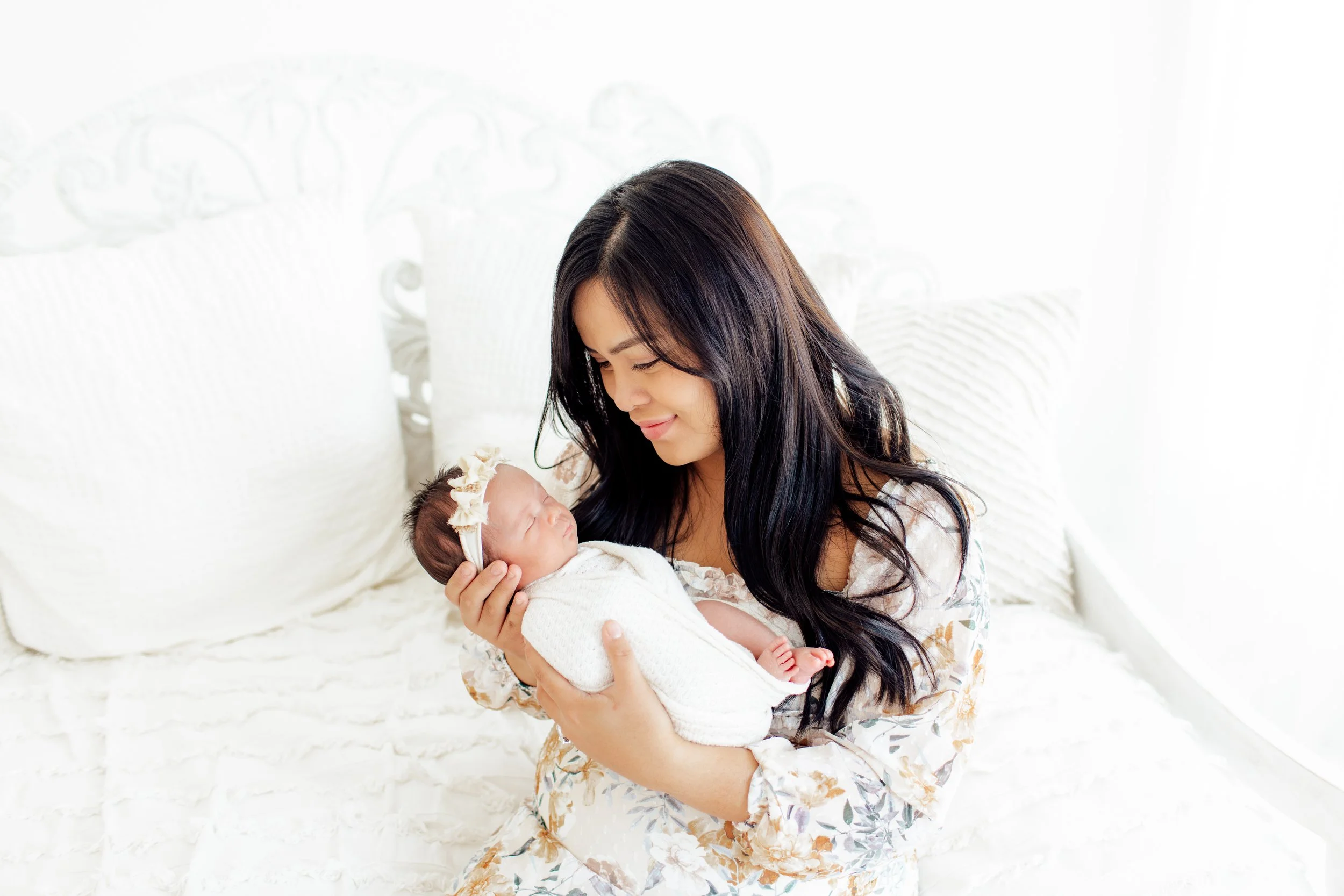 A woman holding a newborn baby on a bed with white bedding, smiling softly and looking at the baby.
