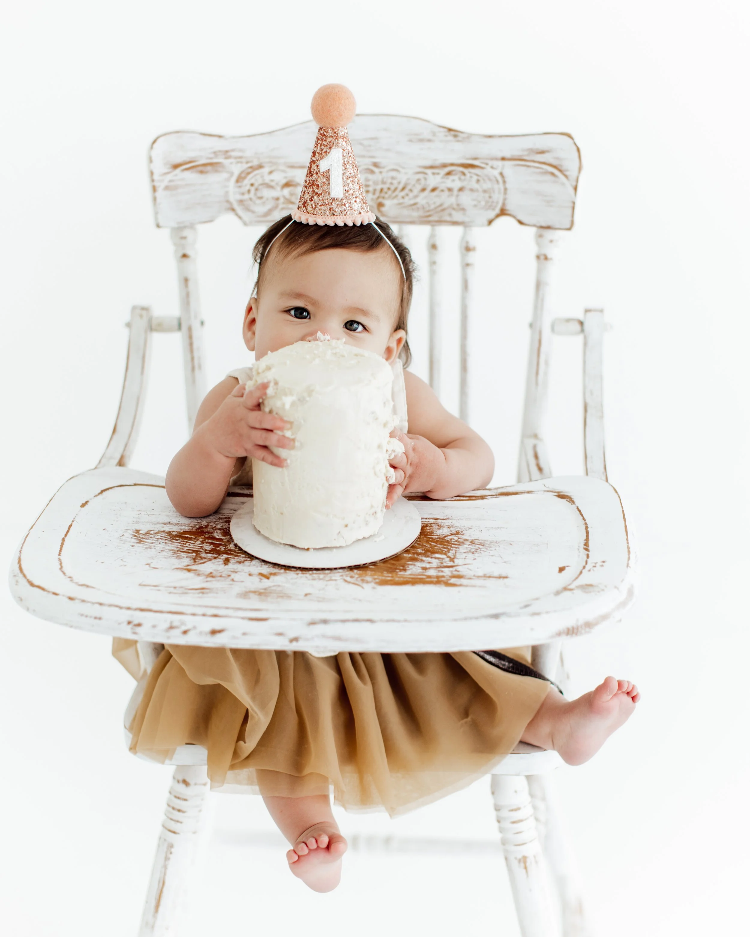 Baby sitting in a high chair, holding a large piece of white cake, wearing a pink birthday hat with a number one, celebrating first birthday.