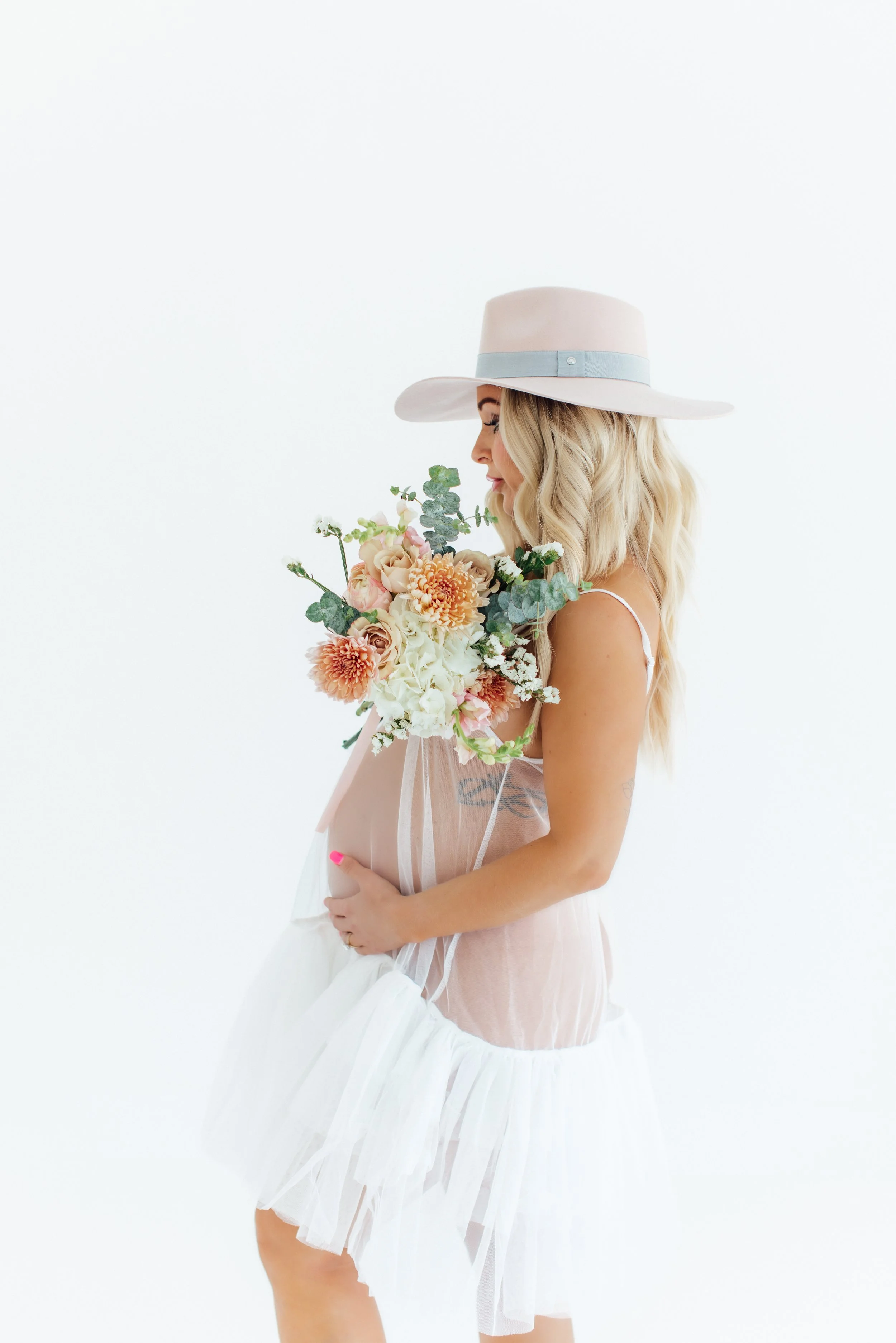 Pregnant woman wearing a wide-brimmed hat, holding a bouquet of flowers, dressed in a sheer, white, ruffled dress, against a plain white background.