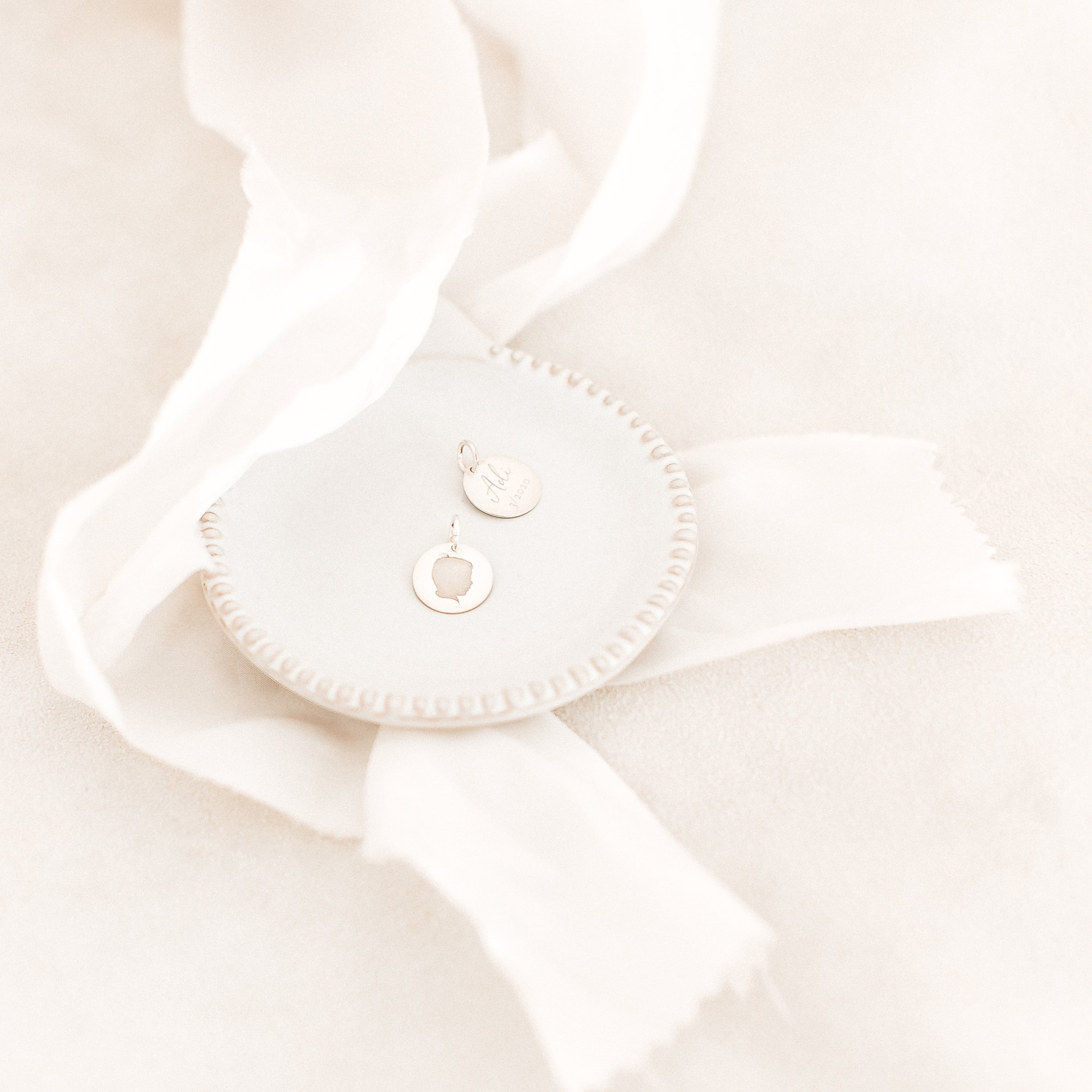 Close-up of two silver charms on a white plate, placed on a white surface with white ribbon and tissue paper.