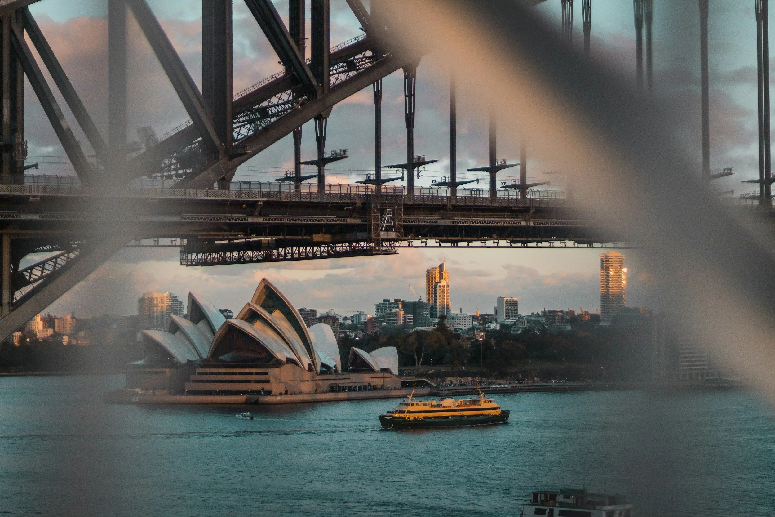 View of Sydney Harbour with the Sydney Opera House in the foreground, a ferry in the water, and the Harbour Bridge overhead, during sunset with nearby skyscrapers in the background.