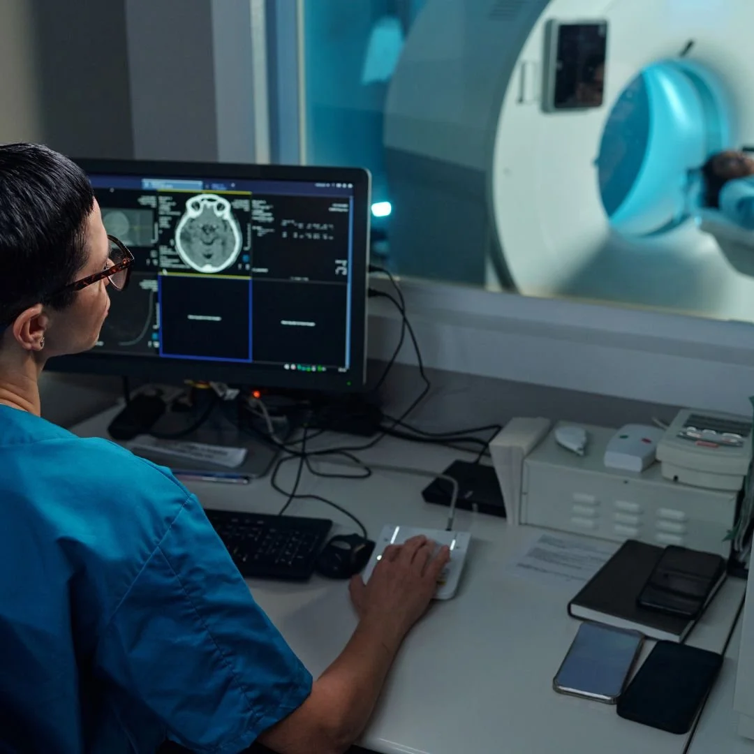 A medical professional monitors a brain scan on a computer screen while operating an MRI machine in a hospital or clinic.