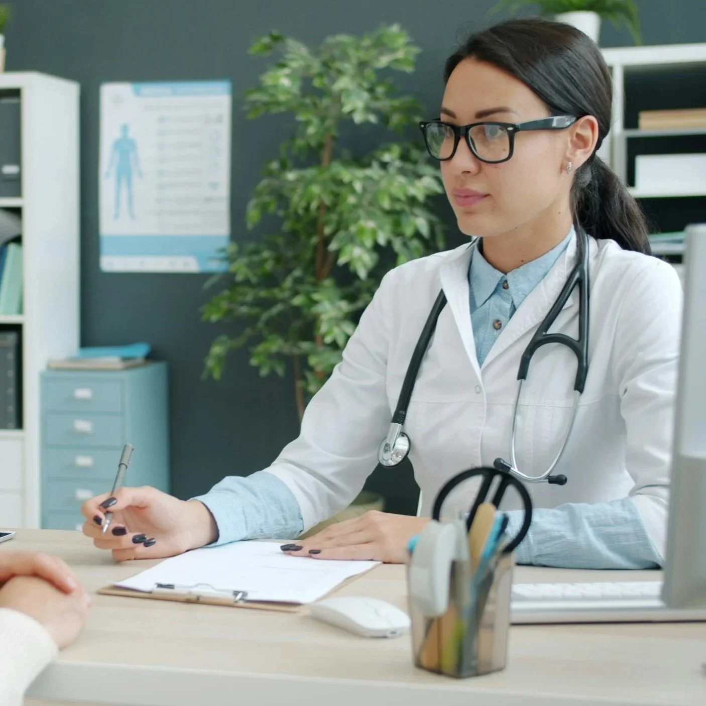 A female doctor with glasses and a stethoscope listens to a patient during a consultation at a medical clinic.