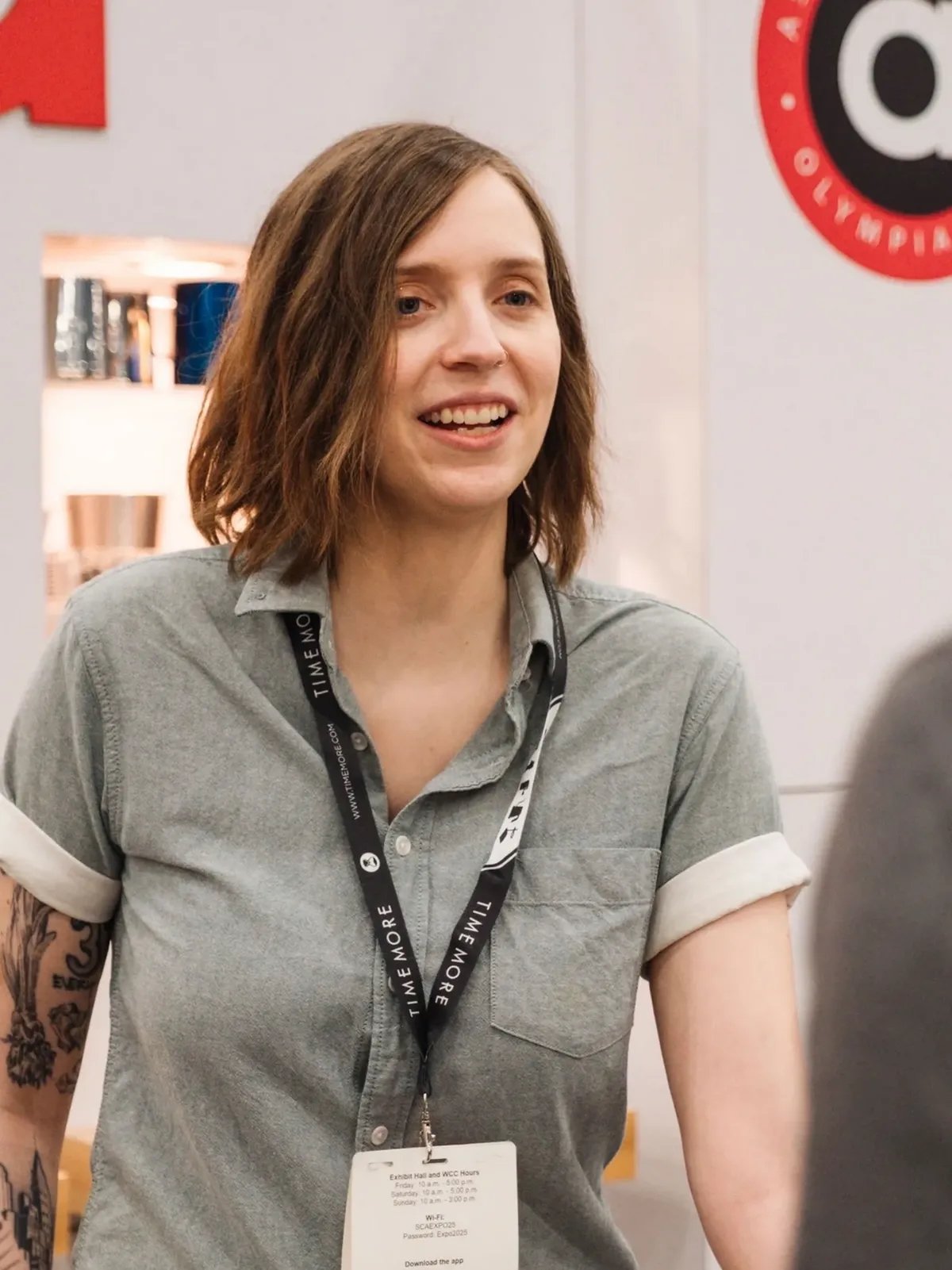 A woman with shoulder-length brown hair, wearing a gray button-up shirt with rolled-up sleeves and a lanyard around her neck, standing indoors and smiling.