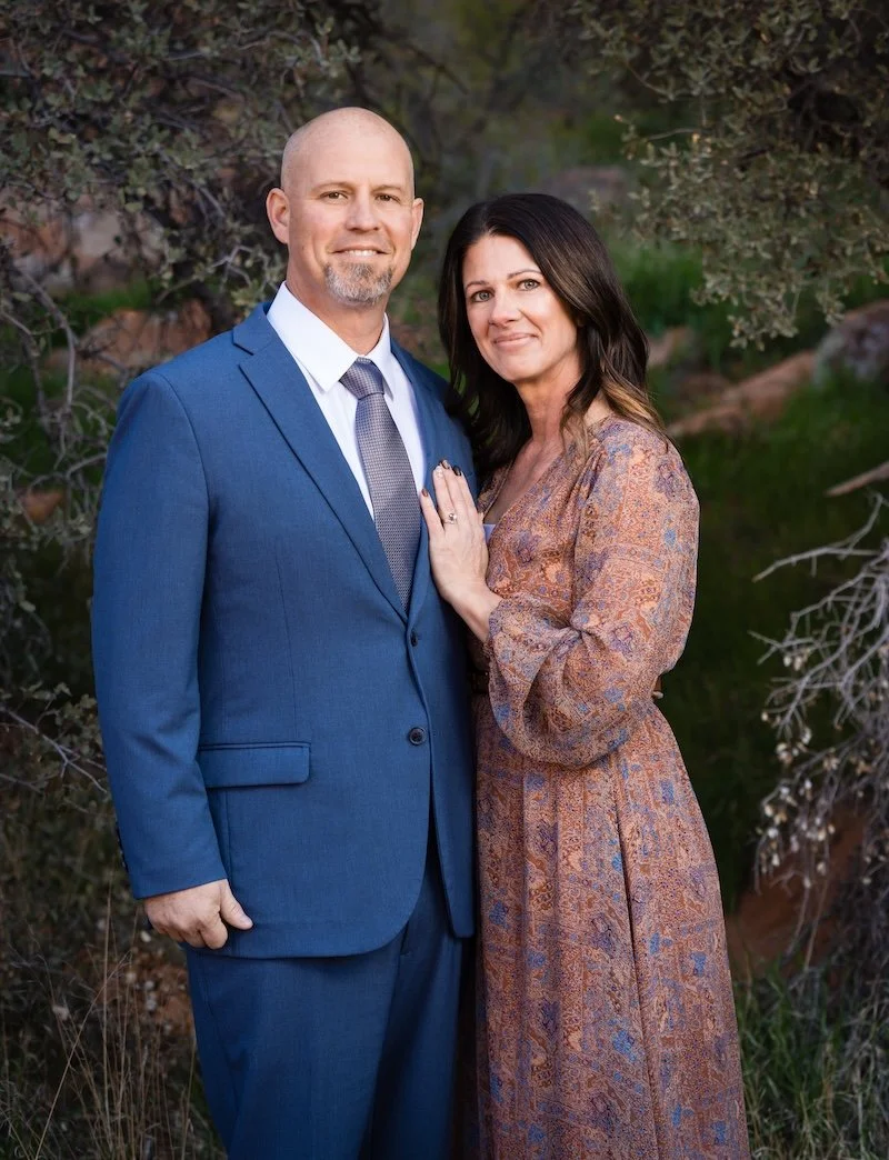 A couple stands outdoors, dressed in formal attire, with the man in a blue suit and tie, and the woman in a patterned long dress, posing for a photo with nature in the background.