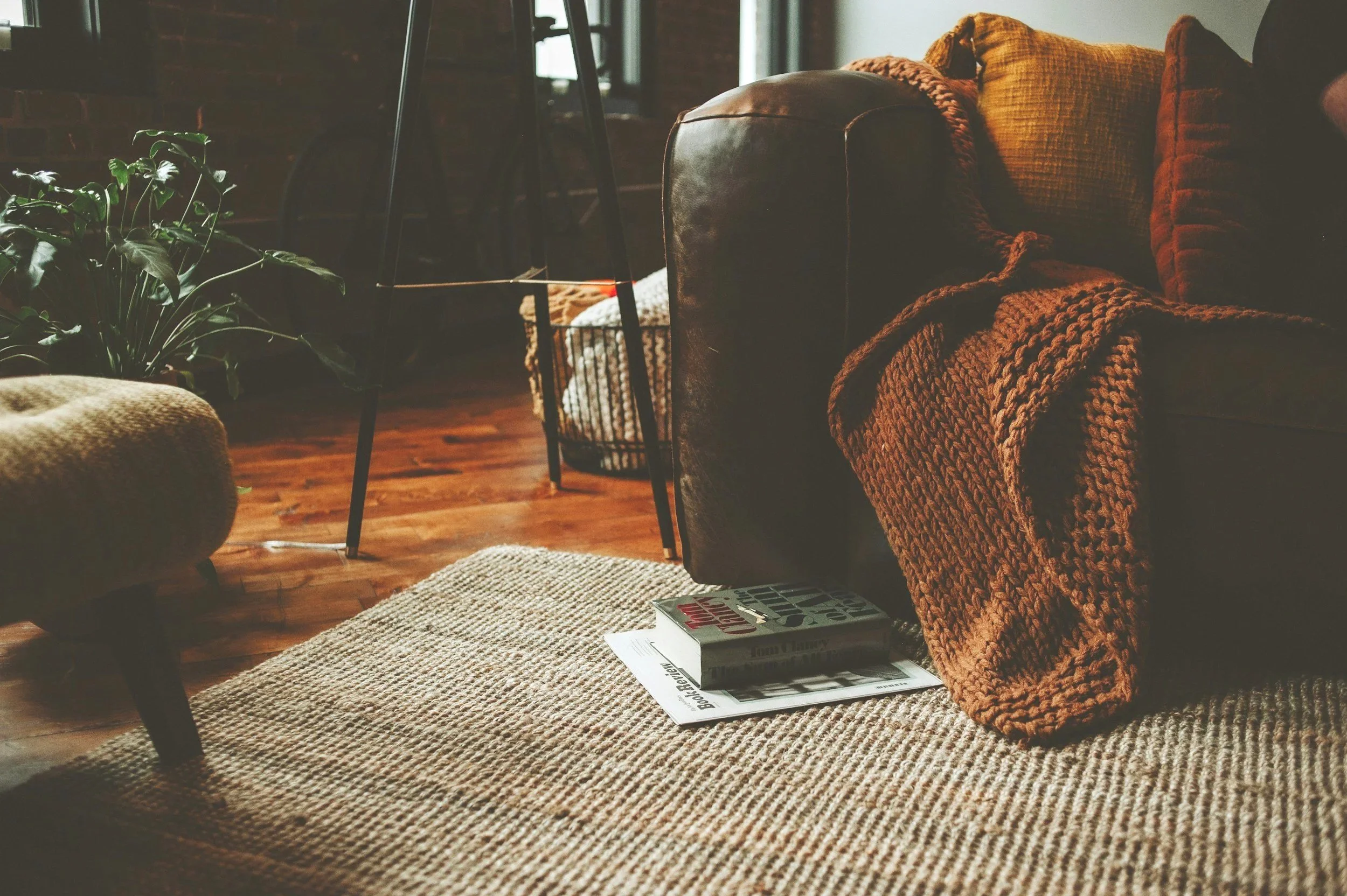 A cozy living room with a woven rug on the wooden floor, a couch draped with a chunky knit blanket, books on the rug, and a potted plant nearby. There's a table with black legs in the background and warm lighting.