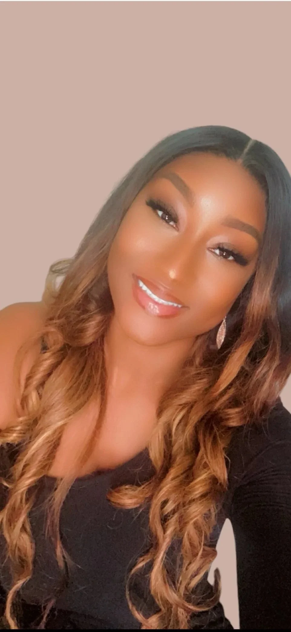 A smiling woman with long, curly hair, wearing a black top and ornate earrings, against a plain background.