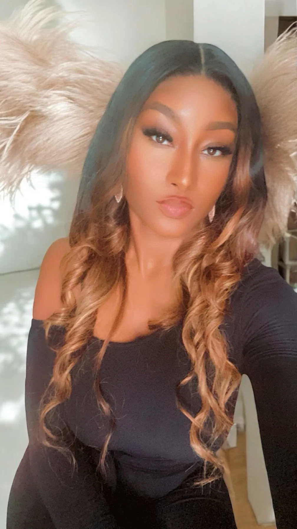 A woman with long, wavy brown and blonde hair taking a selfie. She has makeup on, including eyelashes, and is wearing a black, off-the-shoulder top. Large feathered earrings are visible in her ears, and she is posing confidently.