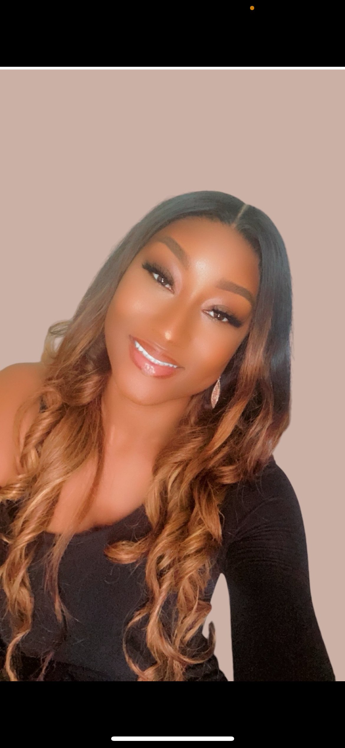 A woman with long curled brown hair and makeup, smiling and wearing a black top and dangling earrings.
