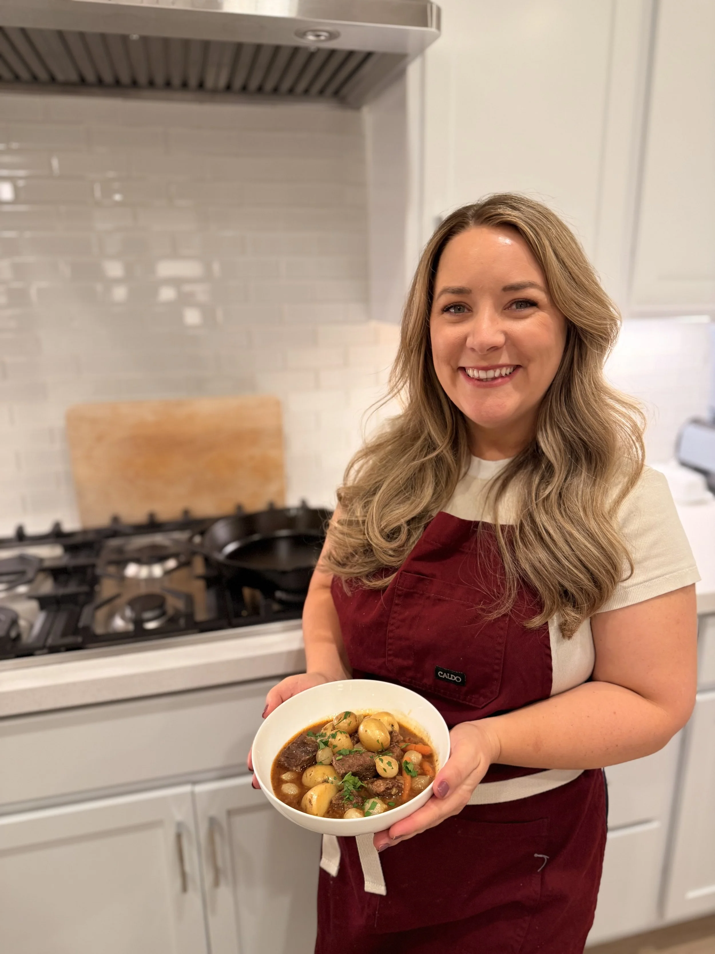 Woman smiling while holding a bowl of beef stew with potatoes and vegetables in a kitchen.