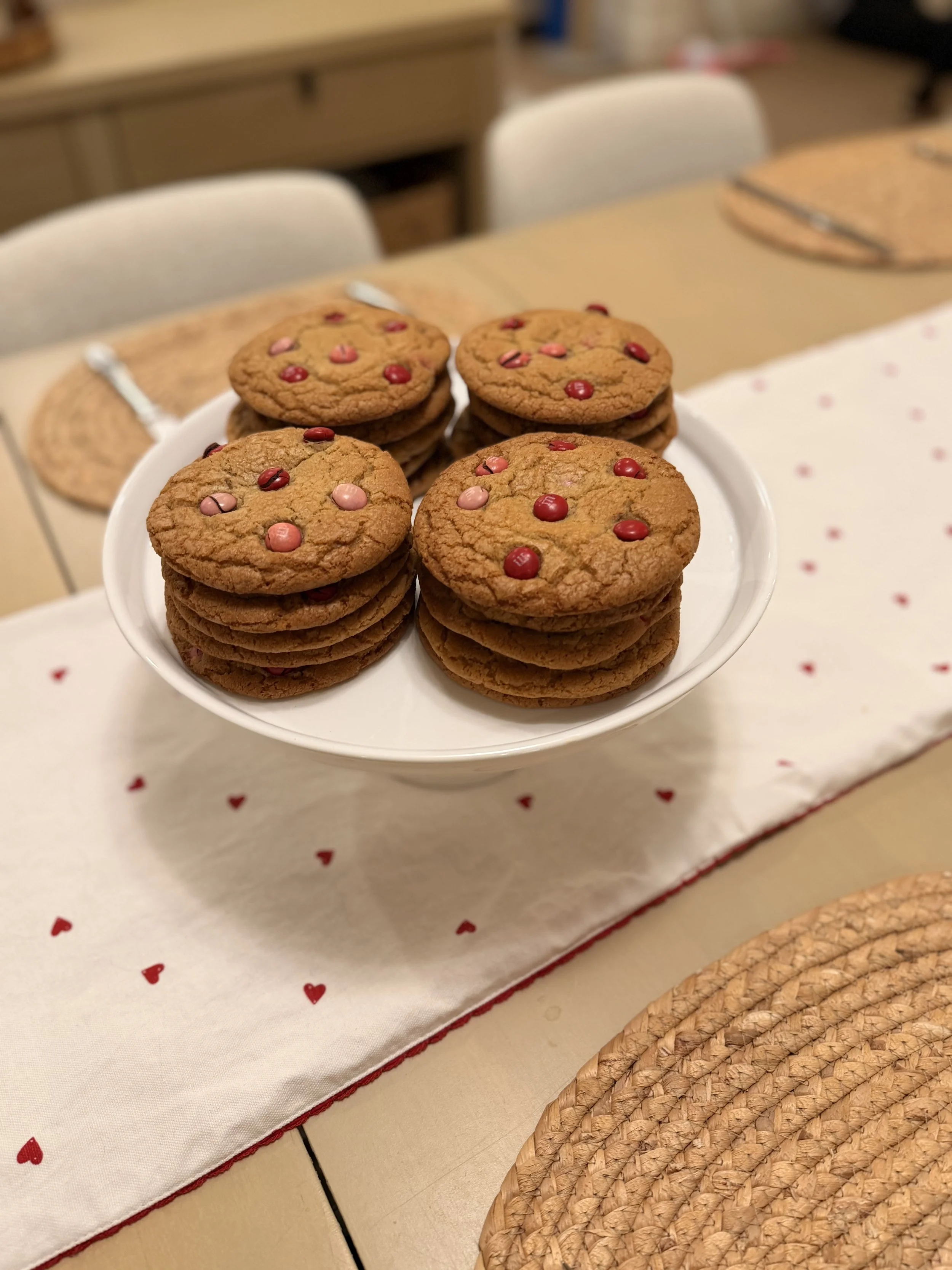 A white plate with M&M cookies decorated with red and pink candy hearts, set on a table with a white table runner with red heart patterns.