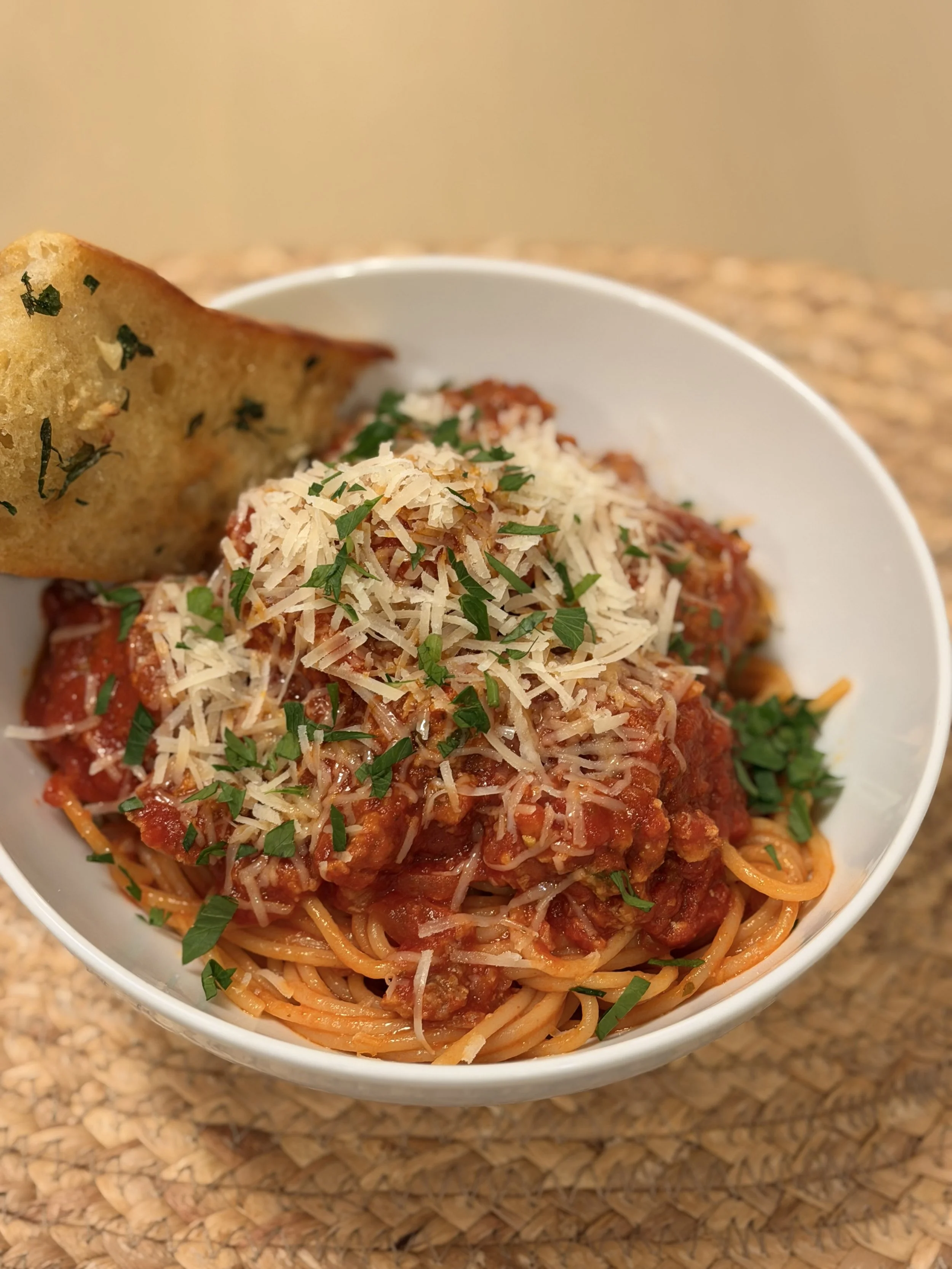 Spaghetti with meat sauce topped with shredded cheese and chopped herbs, served with a piece of garlic bread in a white bowl.