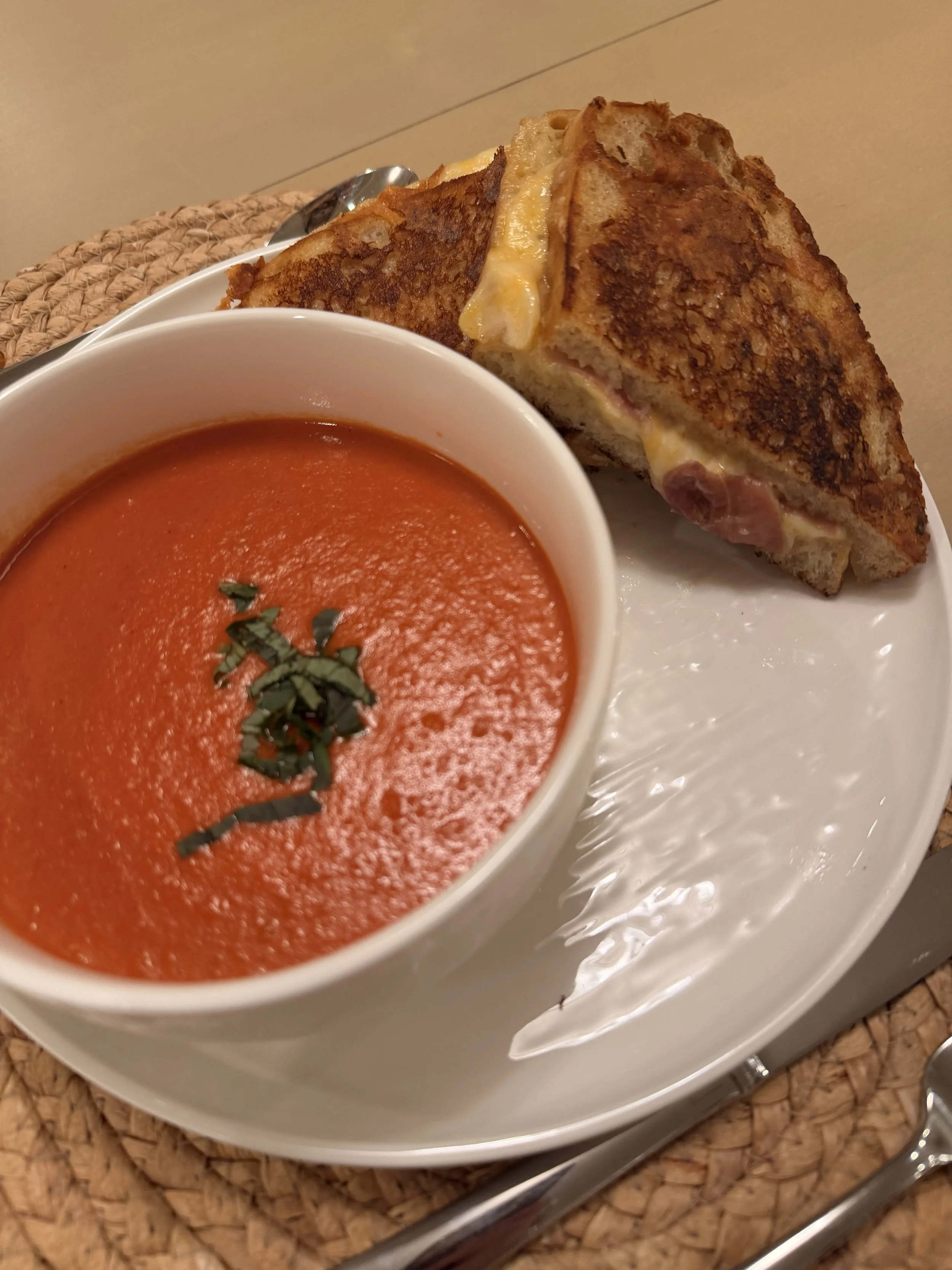 A bowl of tomato soup garnished with basil, served alongside a grilled cheese sandwich on a white plate.