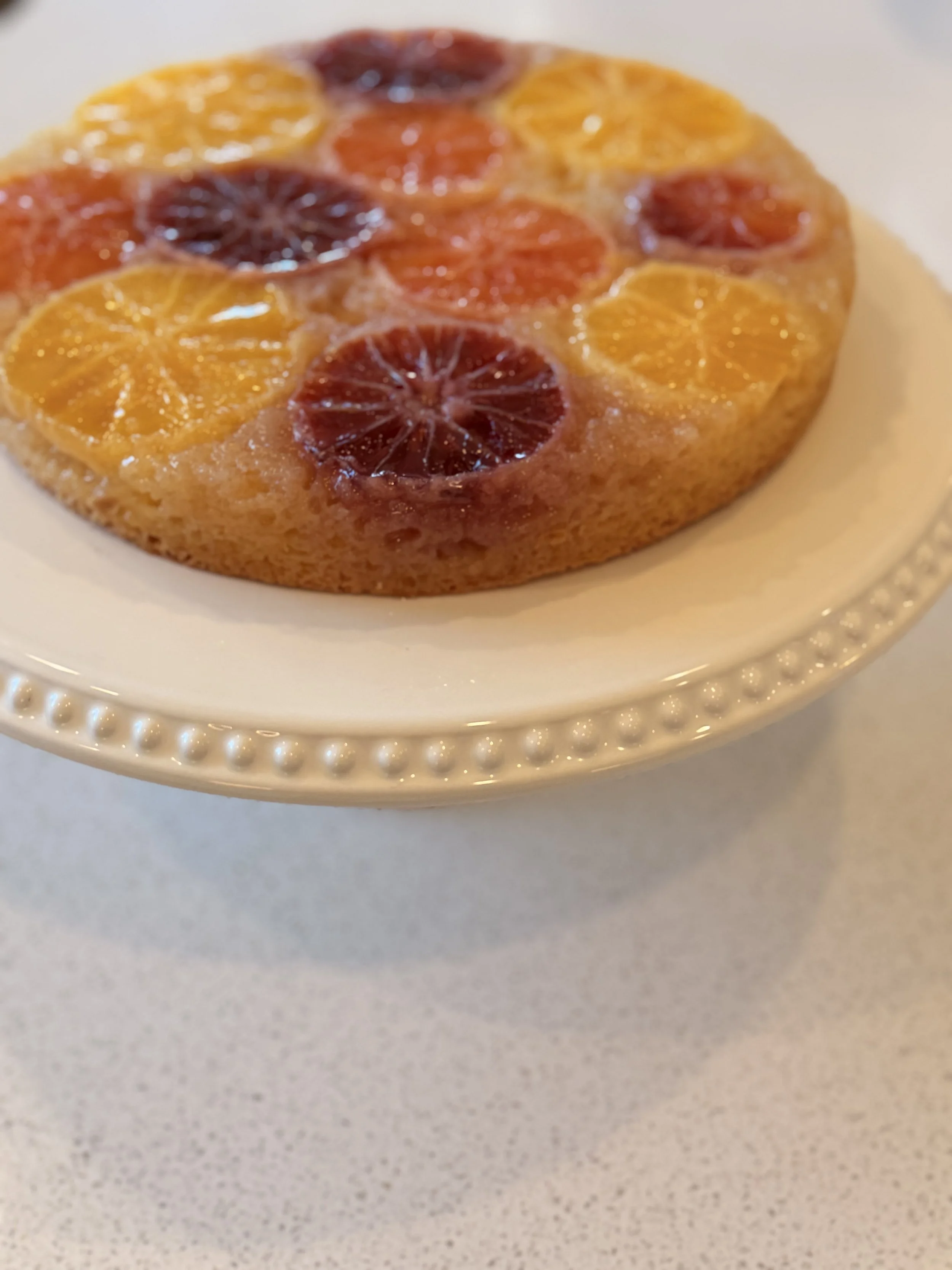 A fruit tart with slices of blood orange and regular orange arranged on top, placed on a white plate with a beaded rim.