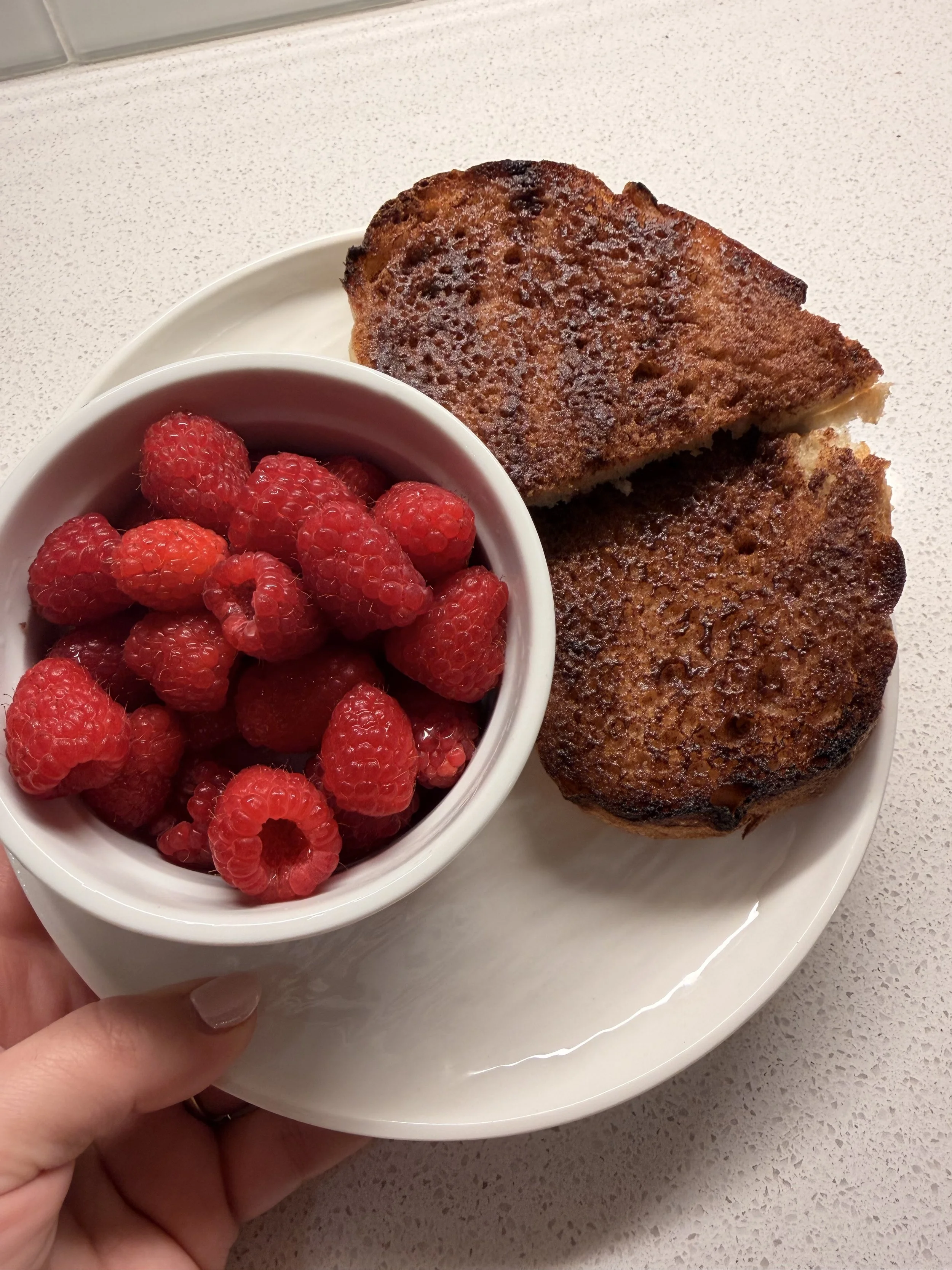 A white plate with two slices of French toast and a small white bowl filled with raspberries, held by a person with a light-colored nail on their finger.