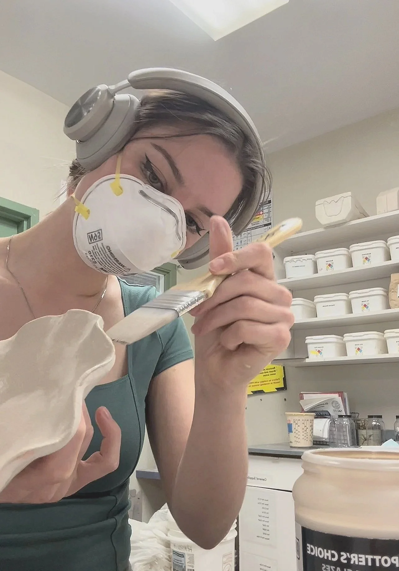 A woman wearing gray headphones and a white face mask is holding a paintbrush and working on a pottery piece in a studio. Shelves behind her are filled with white containers and art supplies.