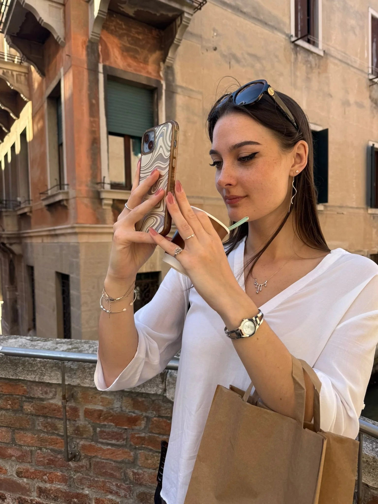 Jazz, wearing sunglasses on her head, earrings, and jewelry, is holding a smartphone with a marbled case, taking a photo. She is standing outdoors against a rustic building in Venice with windows and a brick wall.