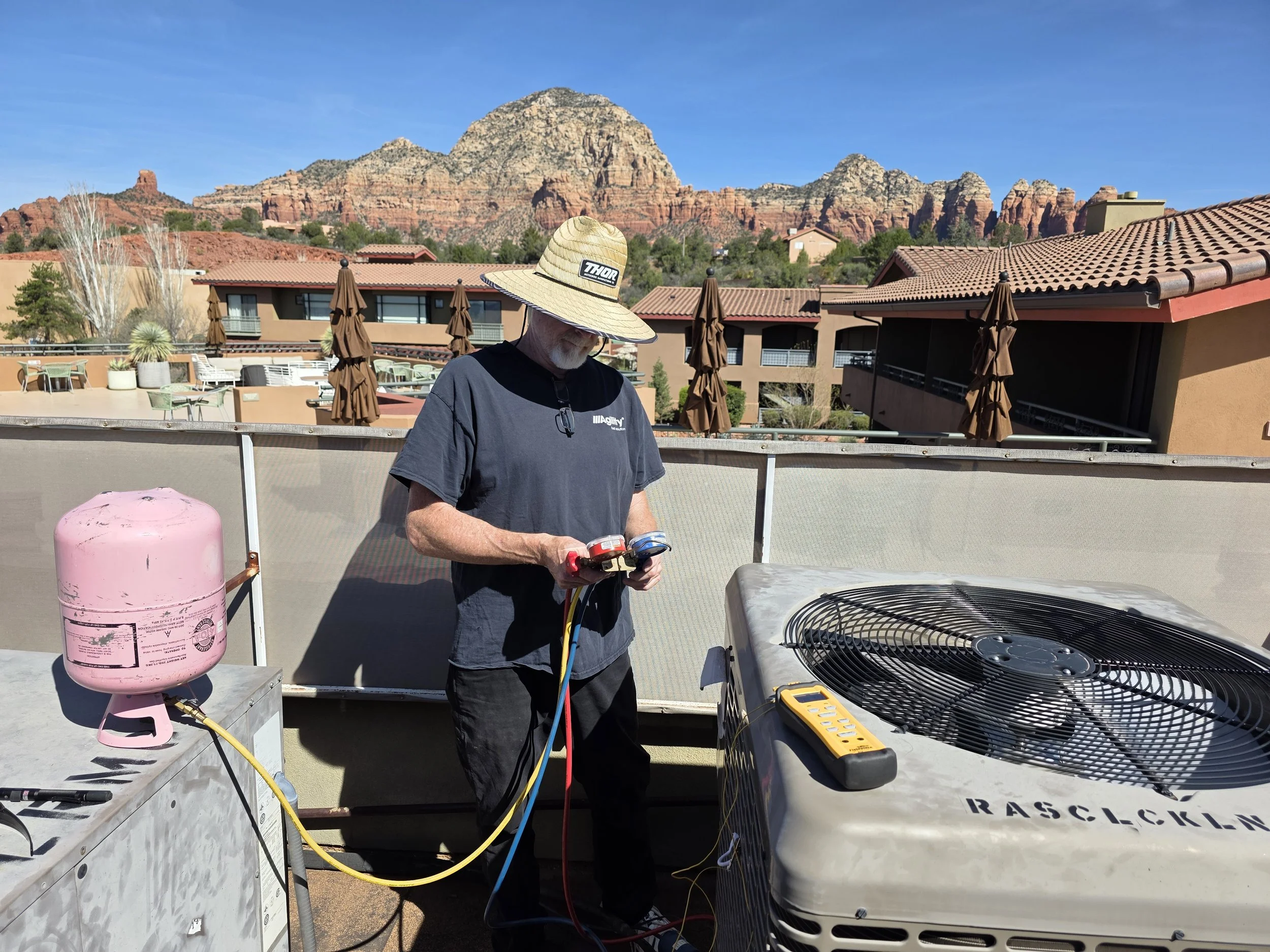 An older man with a wide-brimmed hat working with HVAC equipment on a rooftop, holding a manifold gauge set, with a pink refrigerant tank nearby and a large outdoor fan unit, and red rock mountains in the background under a clear blue sky.
