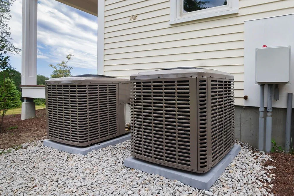 Two large outdoor air conditioning units outside a house on gravel ground.