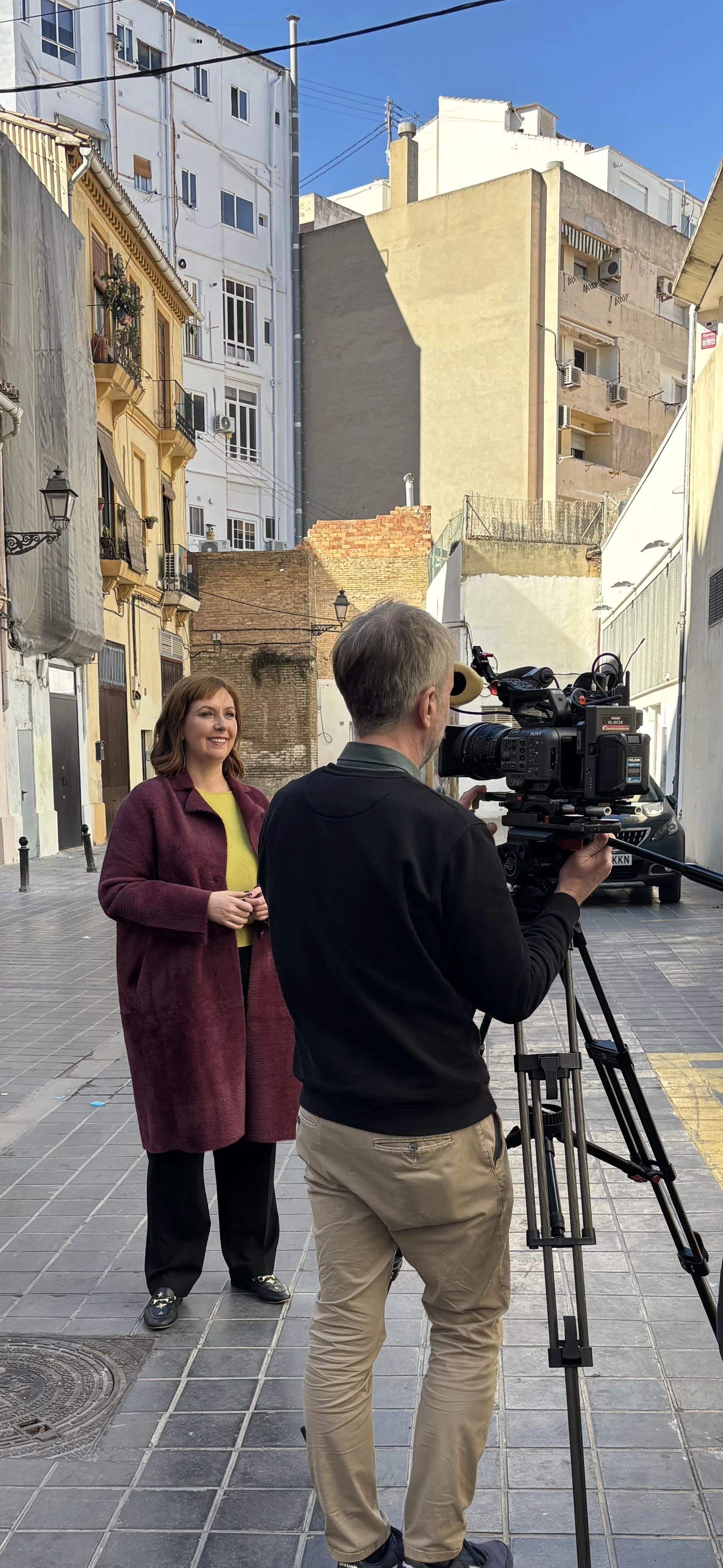 A woman standing in front of a camera being operated by a man on a city street with tall buildings and a clear blue sky.