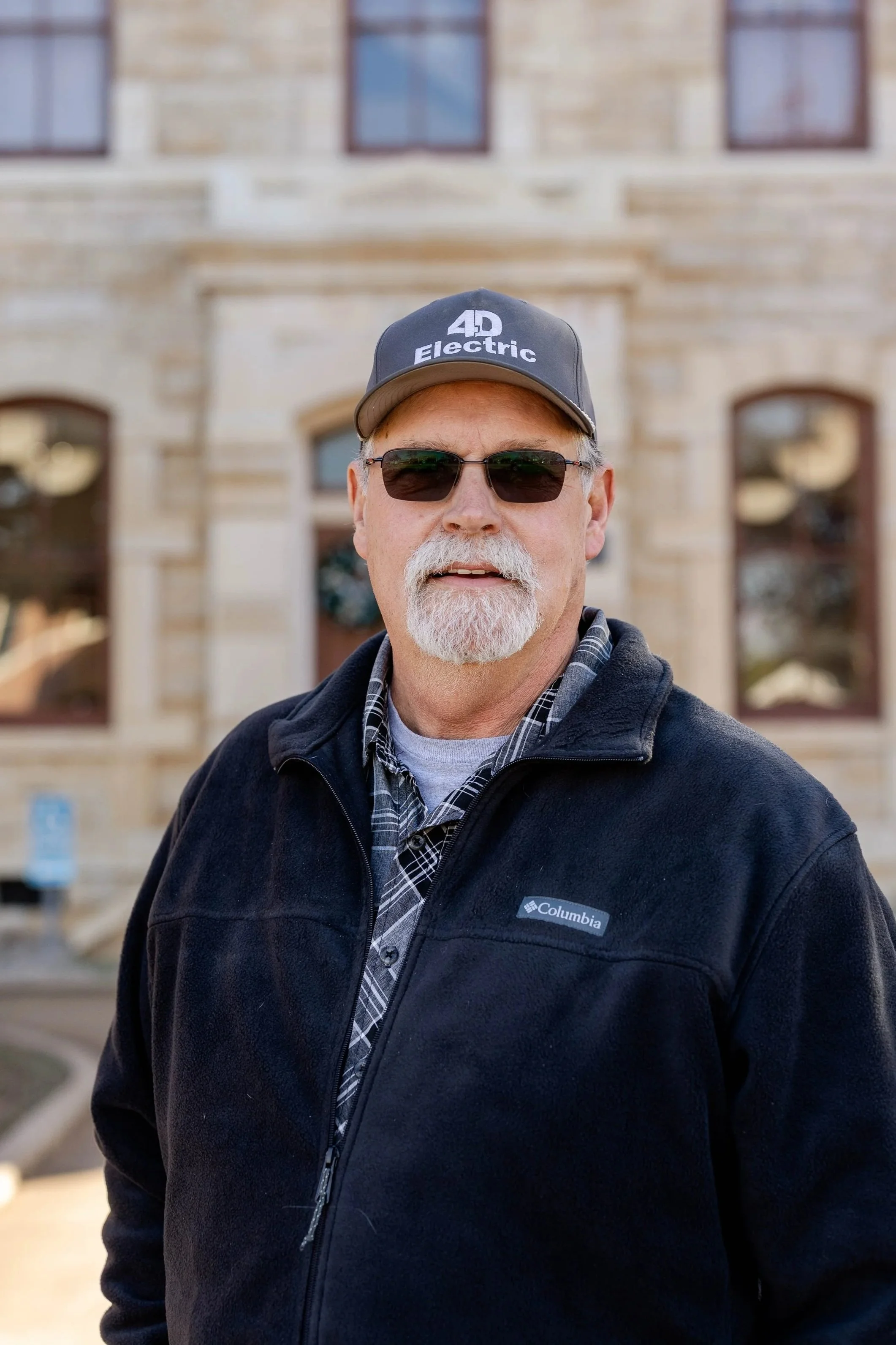 A man with white hair and a beard wearing sunglasses, a baseball cap, and a black Columbia fleece jacket standing outside in front of a stone building.