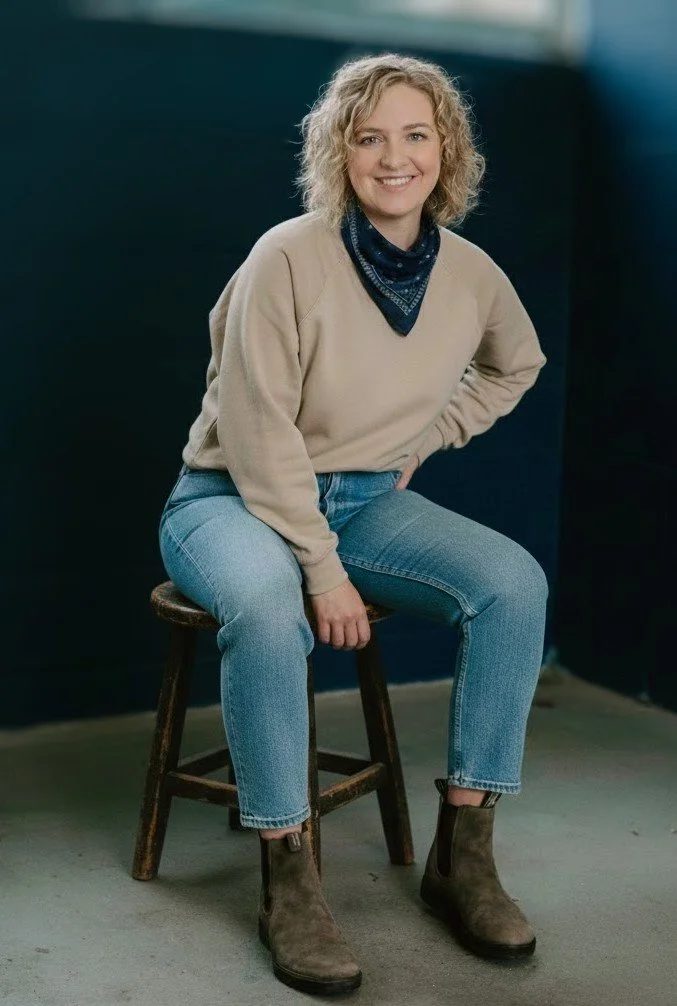 A woman with curly blonde hair, wearing a beige sweater, blue jeans, a navy blue bandana, and brown ankle boots, is sitting on a rustic wooden stool in front of a dark wall.
