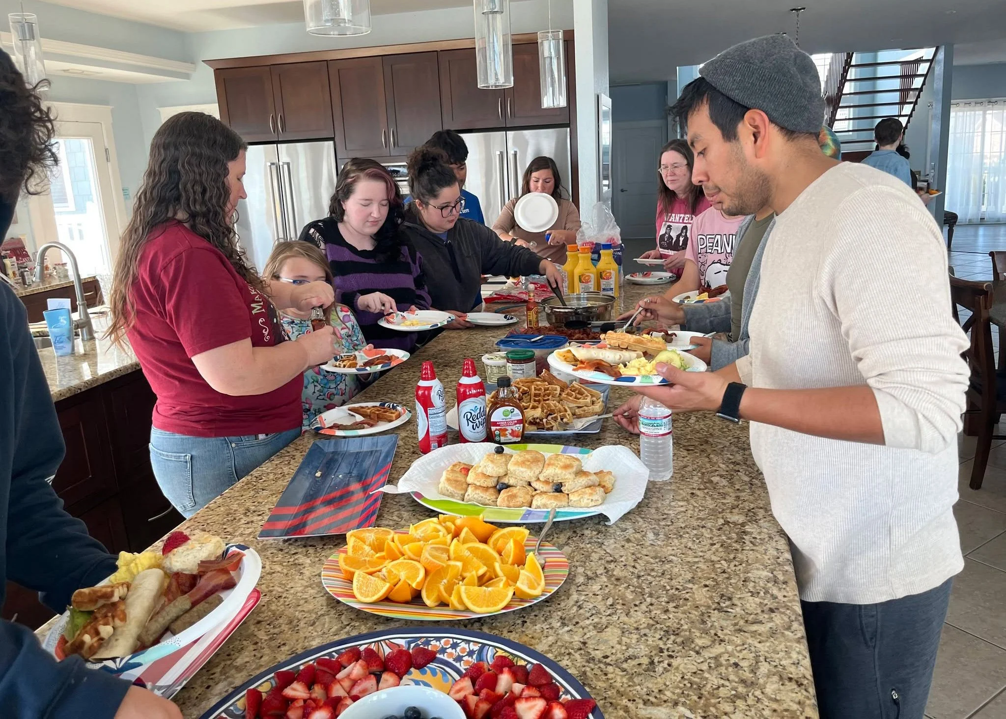 People gather around a kitchen island serving breakfast foods including fruit, waffles, and pastries.