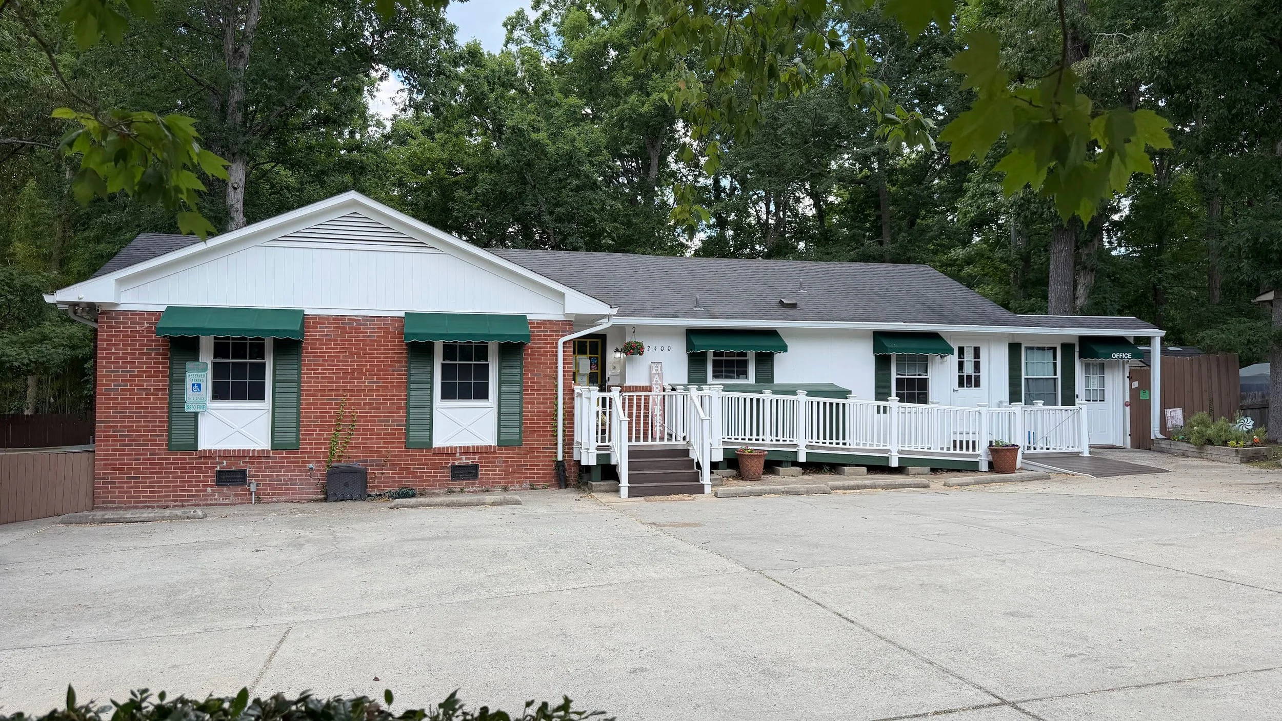 Building with brick and white siding, green shutters, and awnings, a ramp, and outdoor decorations, situated near trees.