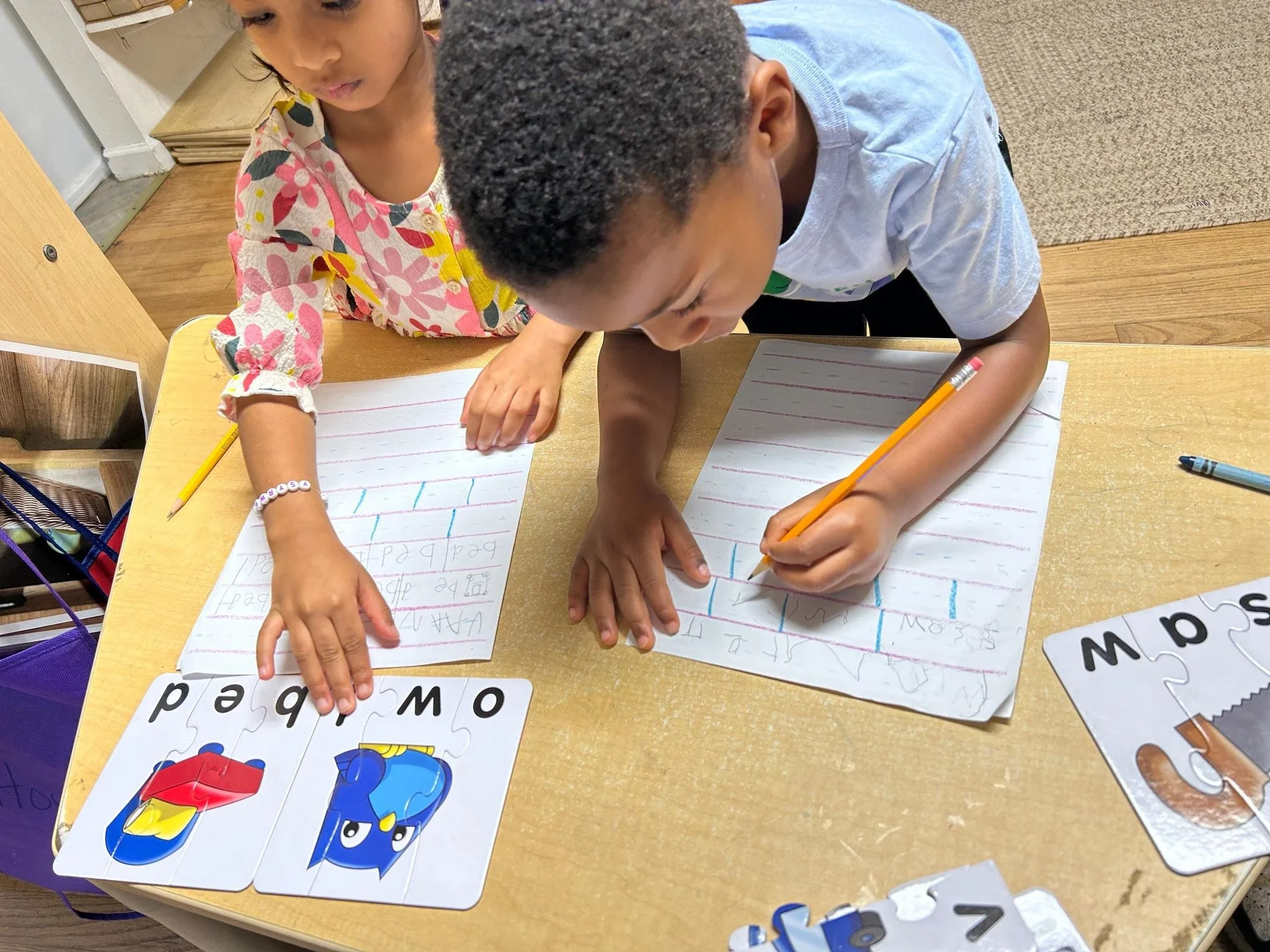 Two children sitting at a table, working on handwriting practice sheets with pencils, while playing with alphabet puzzle pieces.