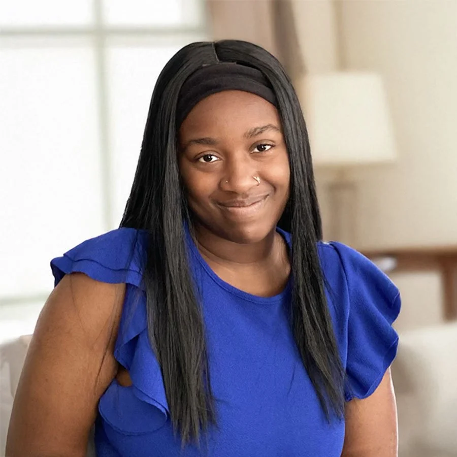 A woman wearing a blue top with ruffled sleeves and a black headband, smiling indoors with a blurred background.