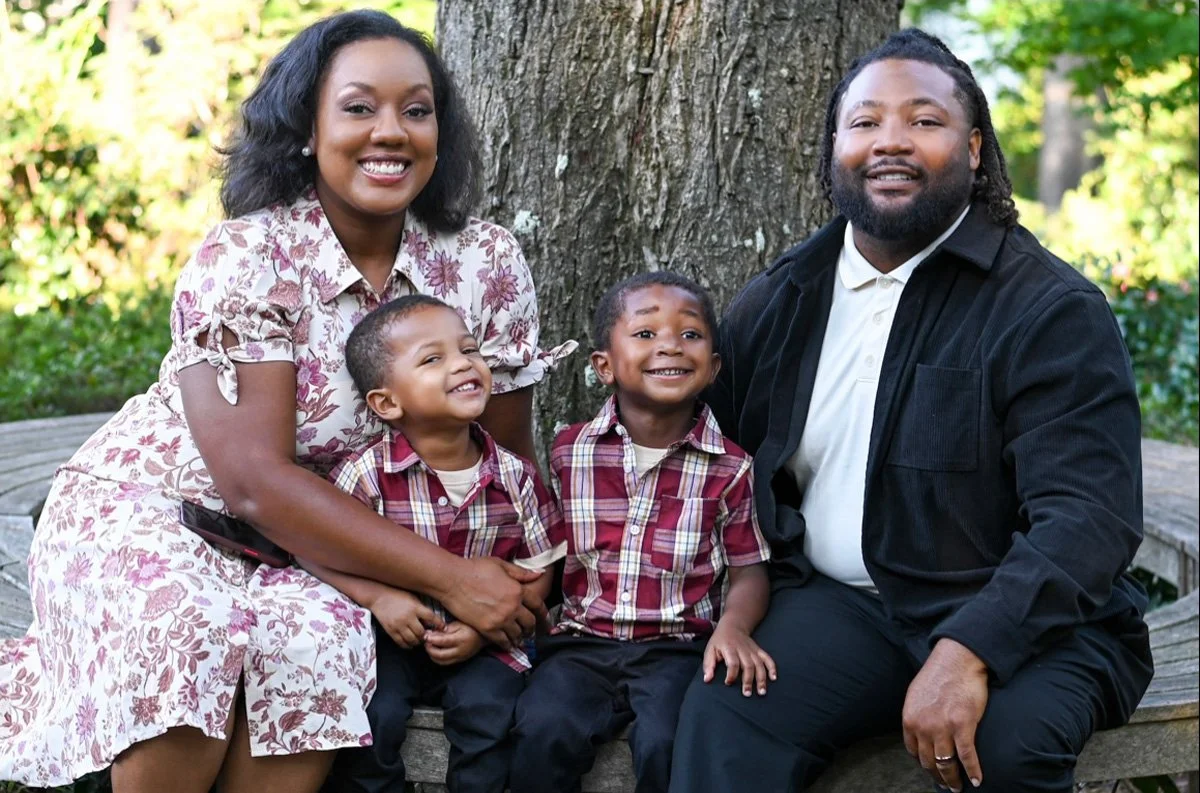 Family of four sitting on a bench outdoors with a tree in the background. The mother and father are with two young boys, all smiling and looking at the camera.
