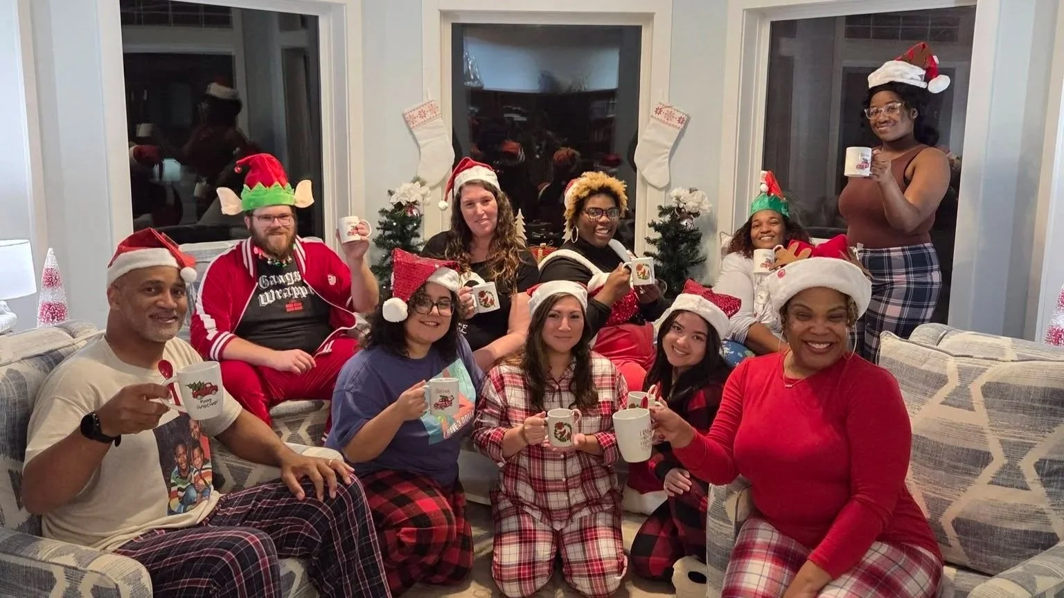 HLC staff are celebrating Christmas in pajamas and holiday hats, sitting on a couch, in a living room decorated for Christmas with stockings and small decorated trees.